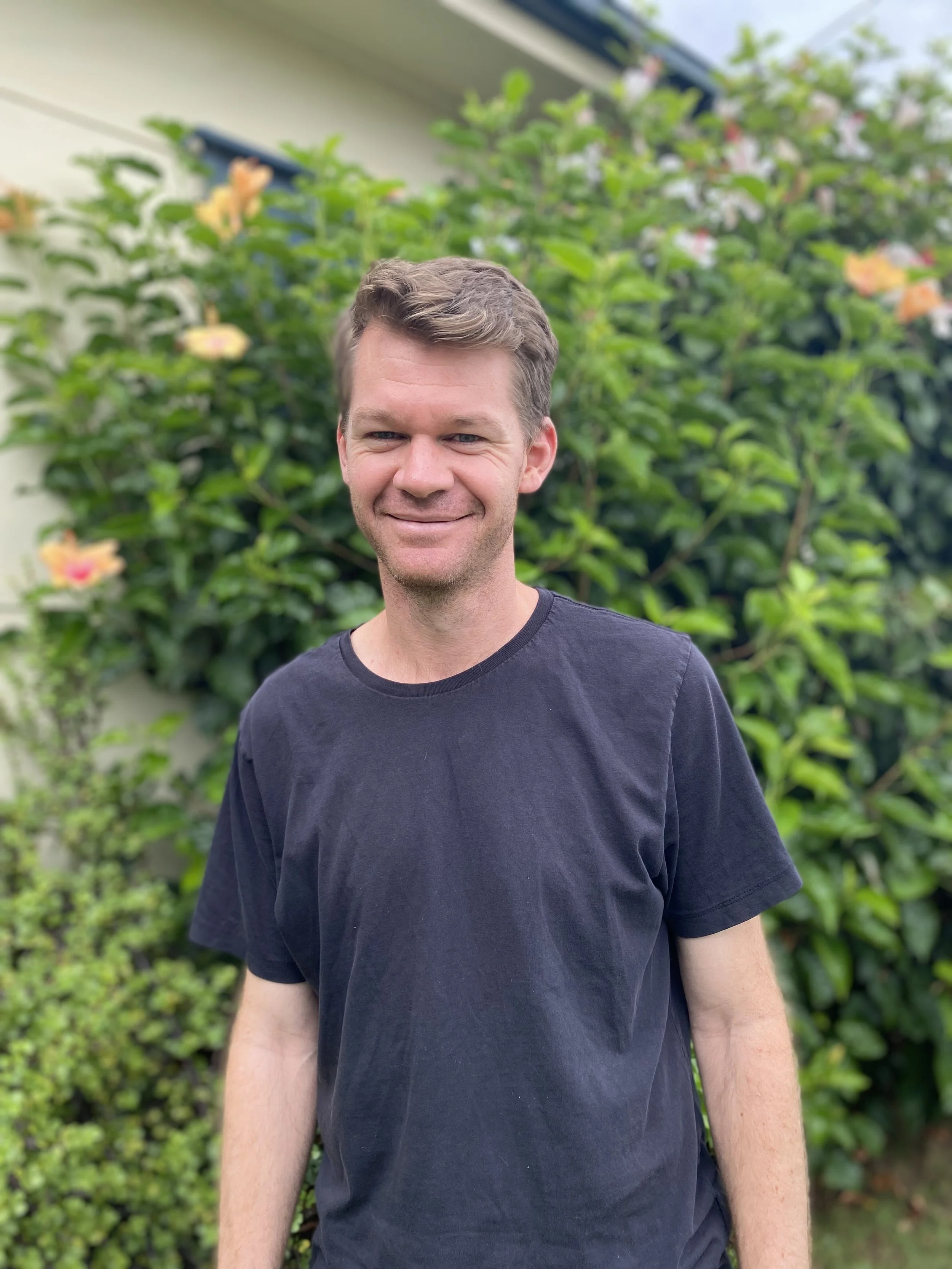 A man with short brown hair and a smile, wearing a black T-shirt, standing outdoors in front of a green bush with pink and orange flowers.