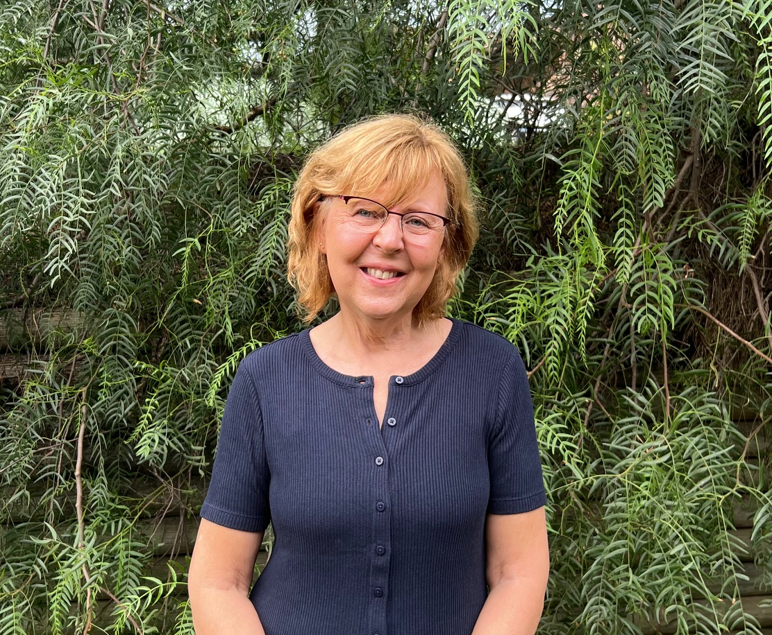 A middle-aged woman with red hair and glasses smiling outdoors in front of green leafy plants.