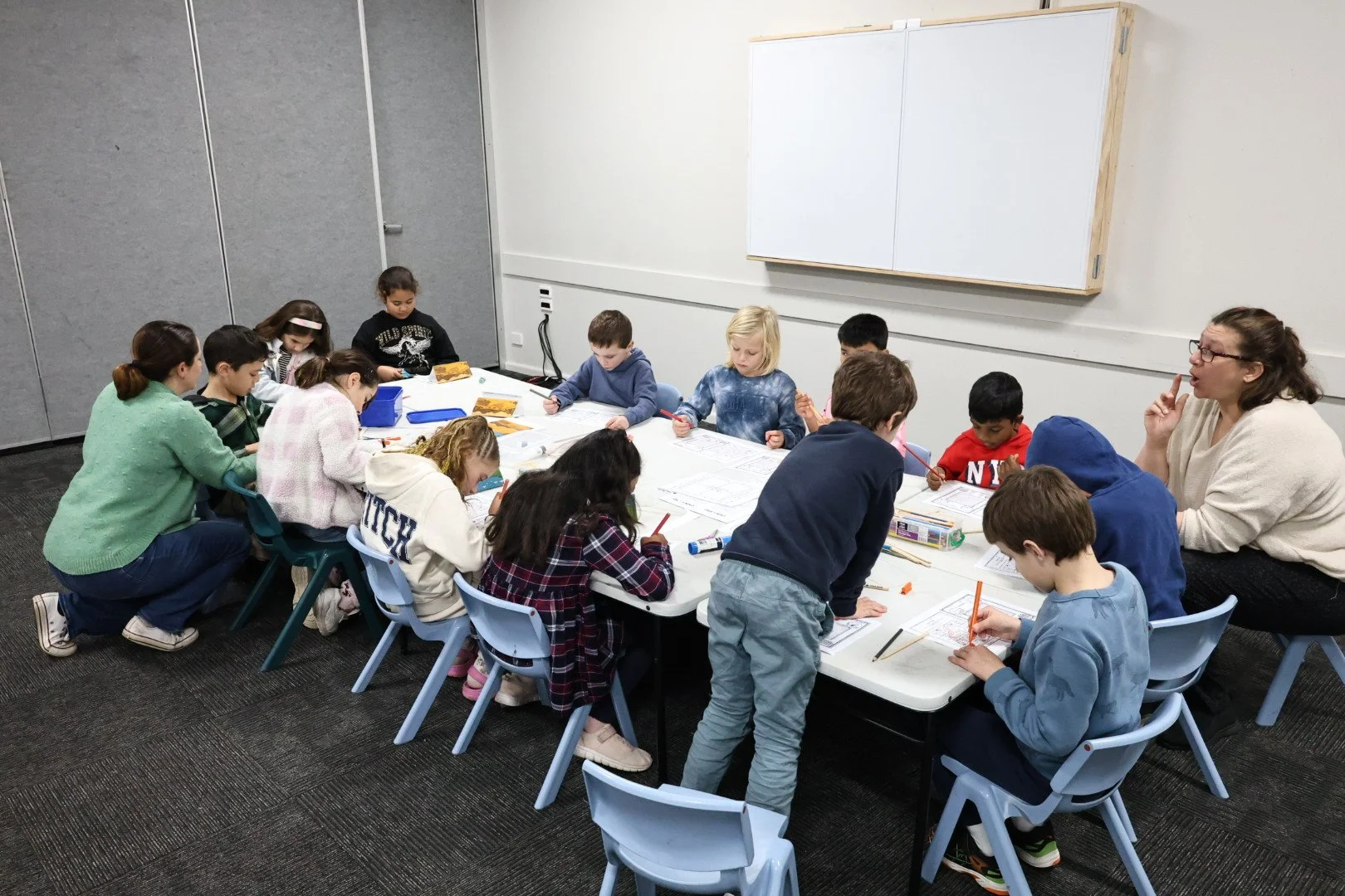Young students sitting around a table in a classroom, engaging in a learning activity with a teacher supervising.