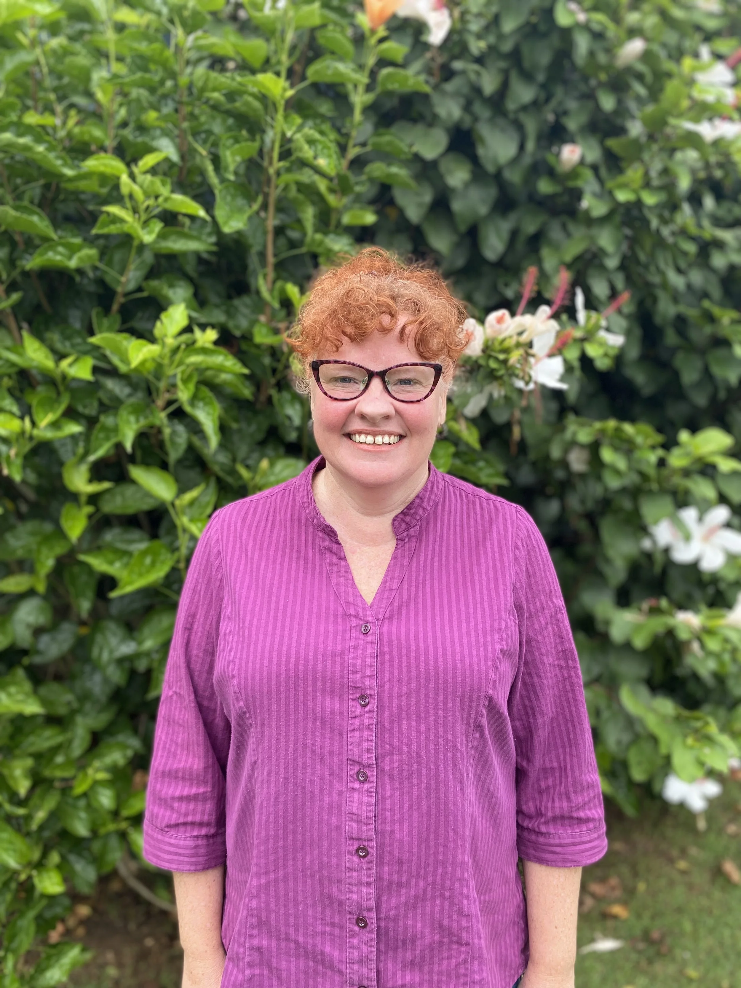A smiling woman with short curly red hair, wearing glasses and a purple button-up shirt, standing outdoors in front of green foliage and white flowers.