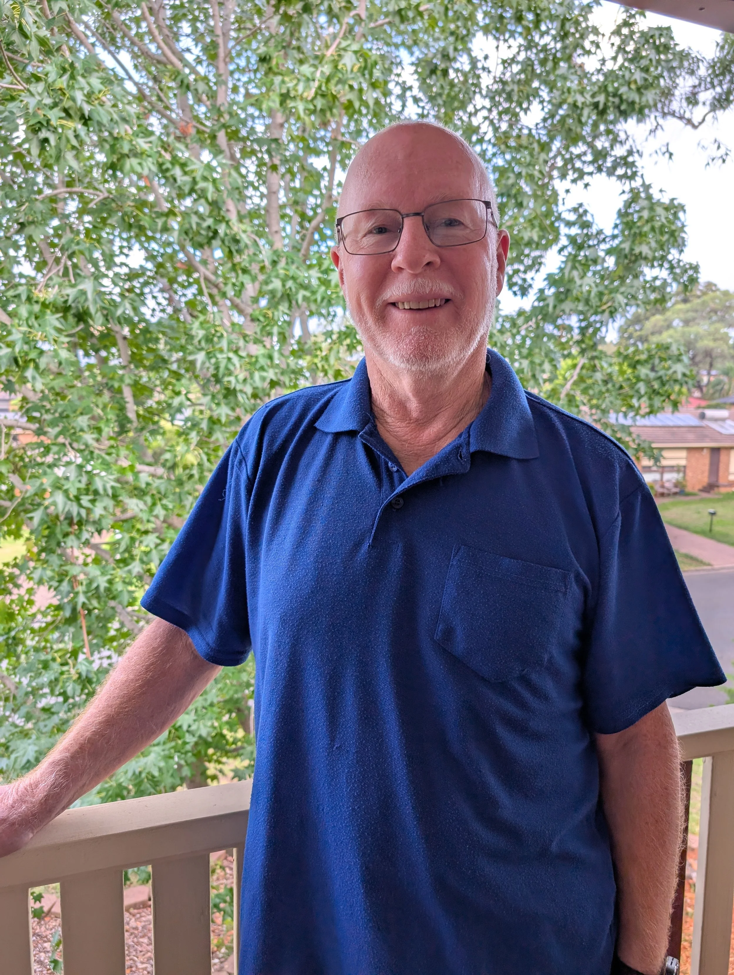 An older man with glasses, a beard, and a bald head, smiling in front of a leafy green tree and a suburban neighborhood, wearing a navy blue polo shirt.