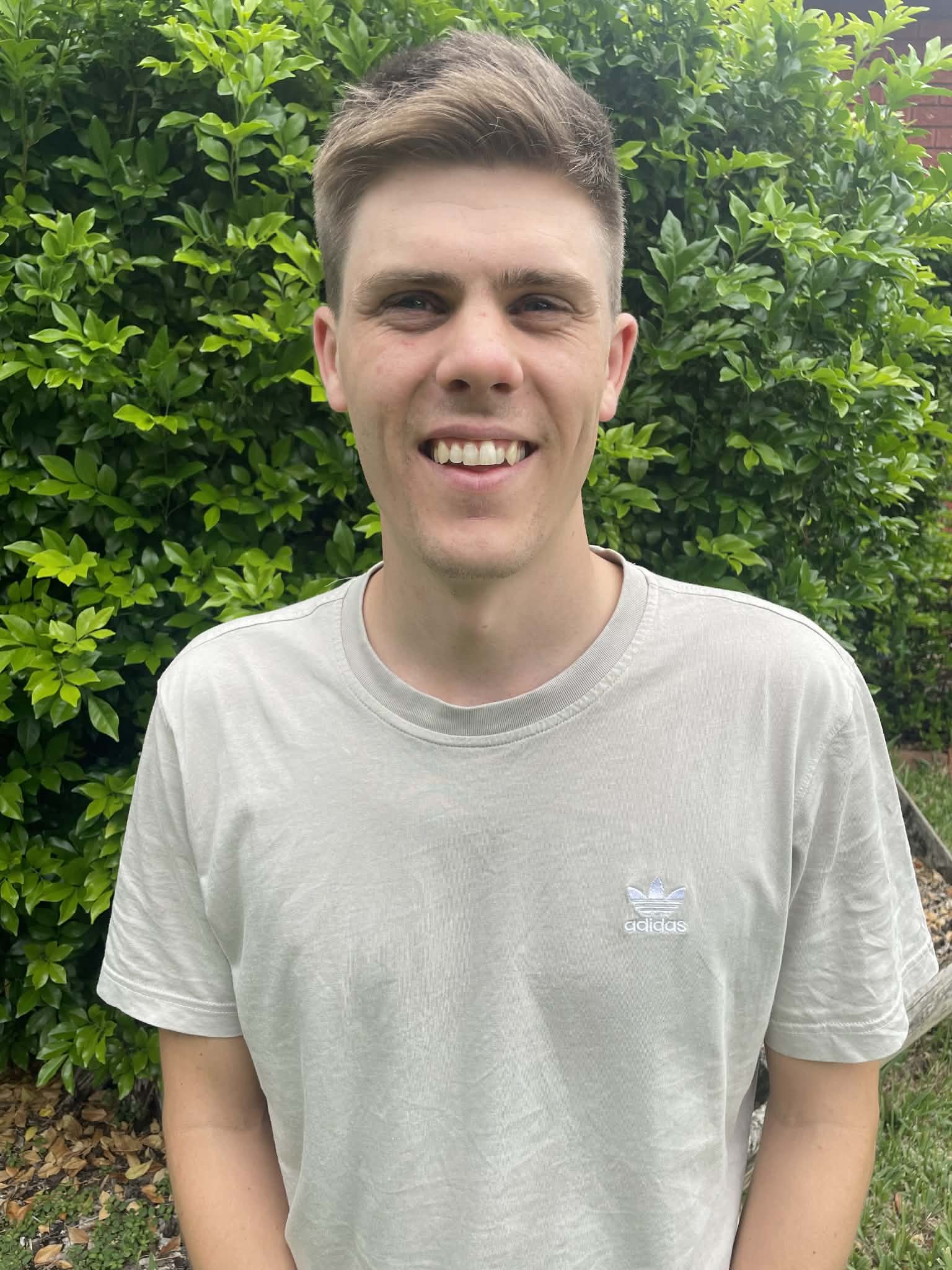 A young man with light skin and brown hair, smiling and standing outdoors in front of a lush green bush, wearing a light grey Adidas t-shirt.