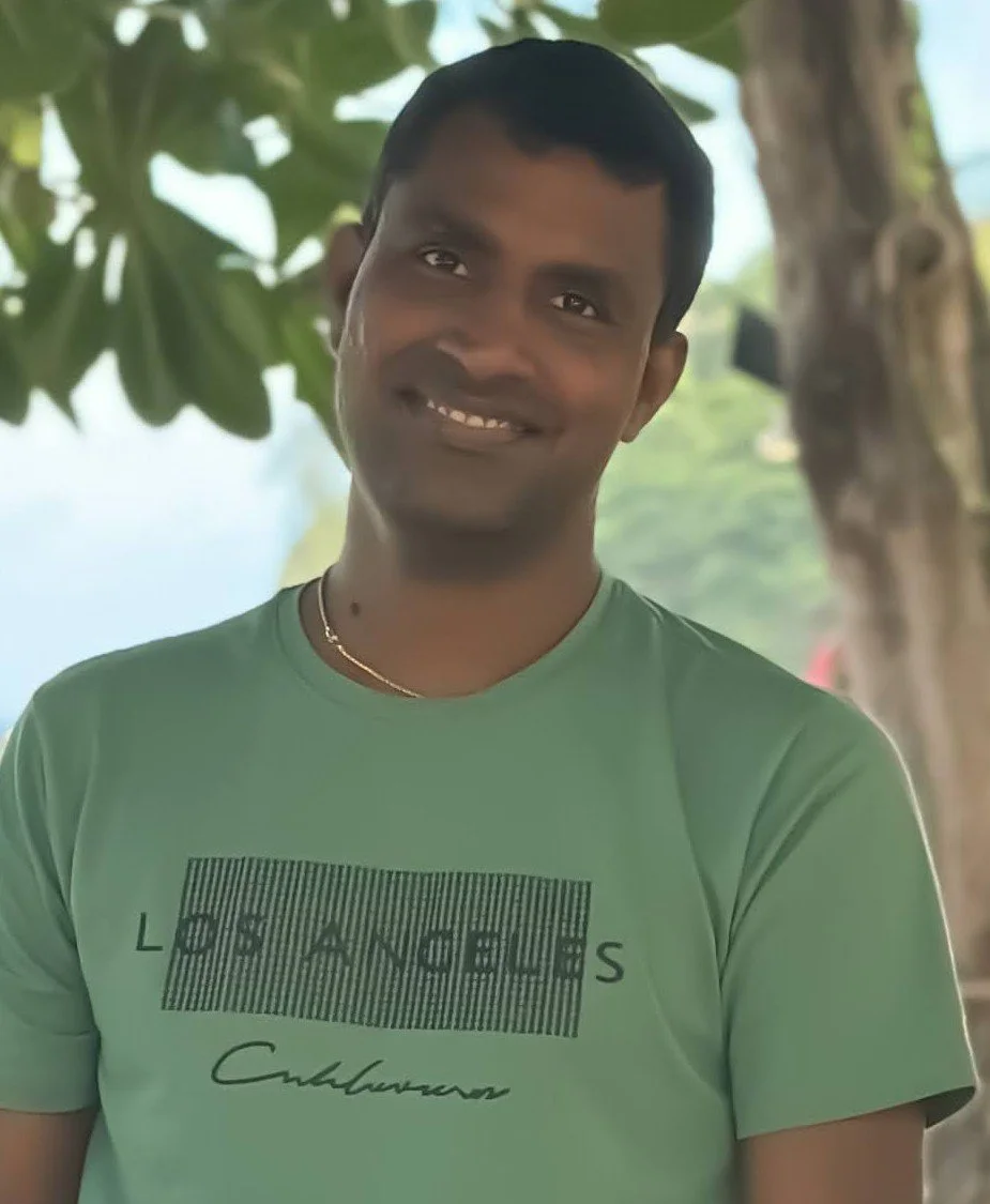 A man smiling outdoors with trees and blue sky in the background, wearing a light green t-shirt with partially obscured text and a gold necklace.