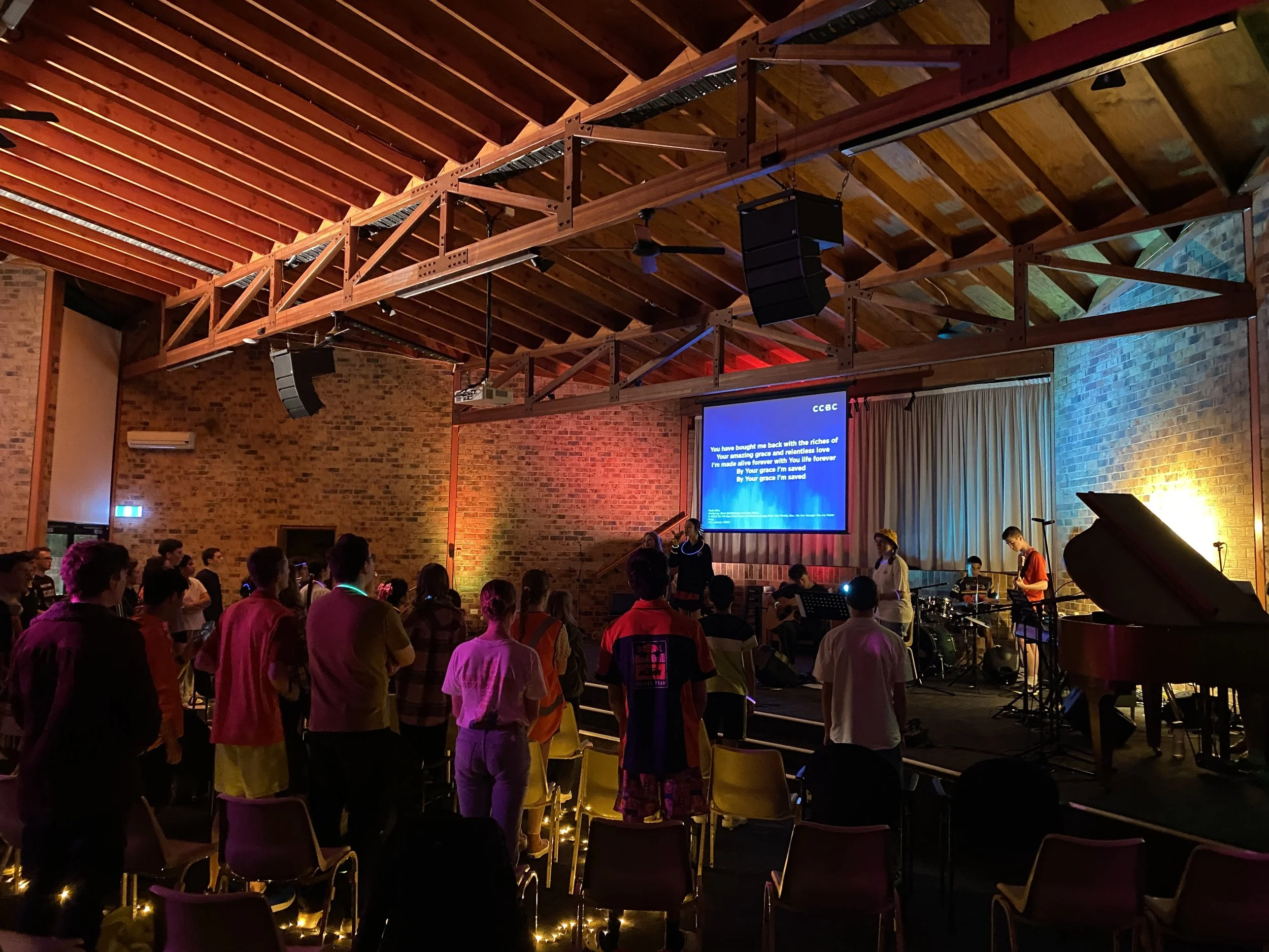 A group of people gathered on a stage in a church or concert hall, with musicians and singers performing in front of a large screen with song lyrics, brick walls, wooden ceiling beams, and colorful stage lighting.