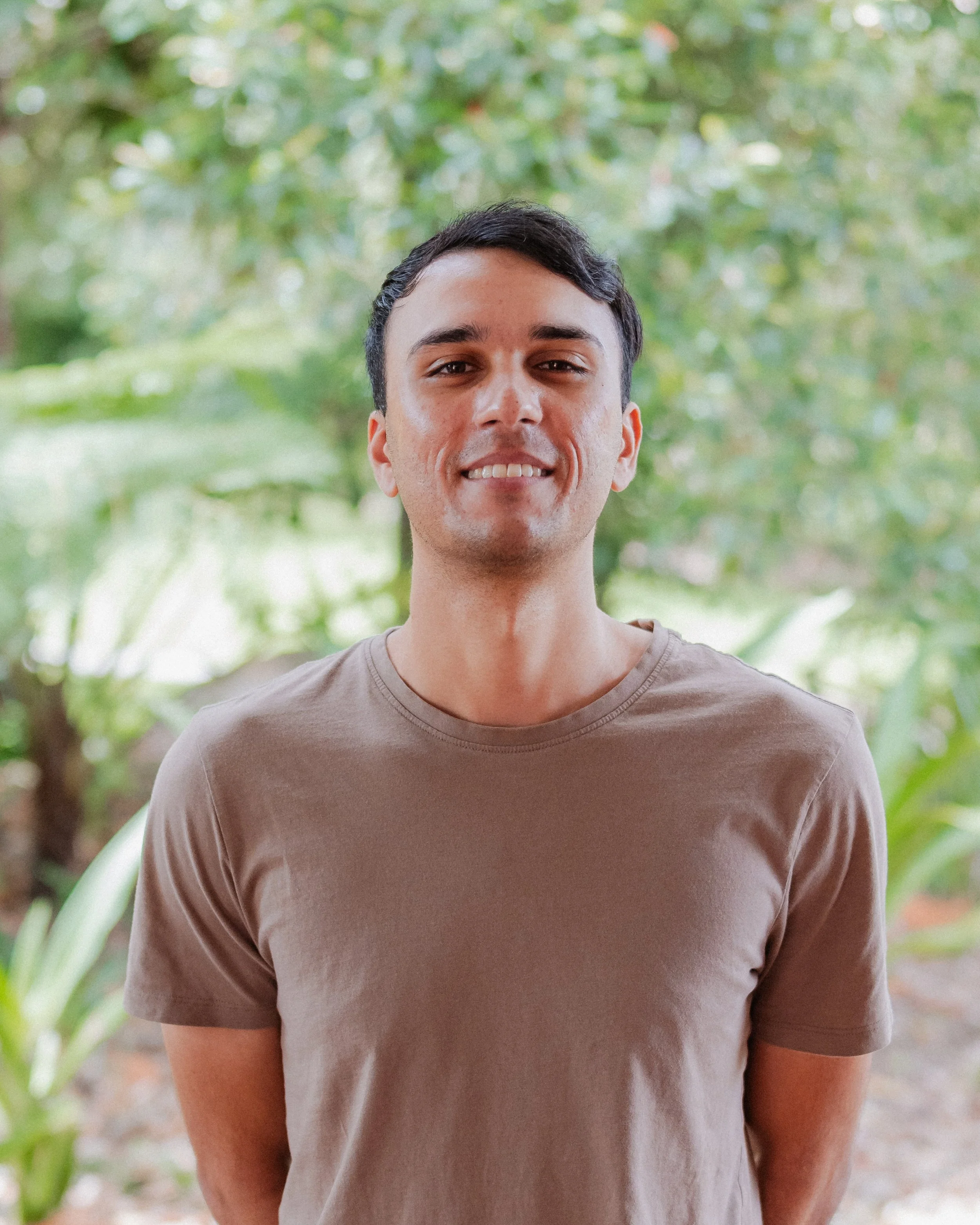A young man with short dark hair, light skin, wearing a beige T-shirt, standing outdoors with plants and trees in the background.