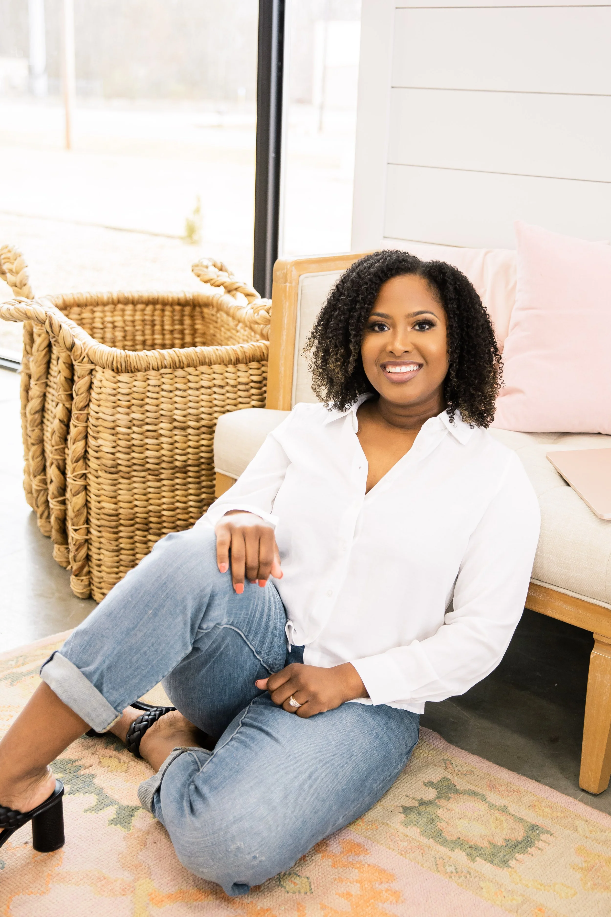 A woman sitting on a rug, smiling, in a cozy indoor setting with wicker baskets and a large window behind her.