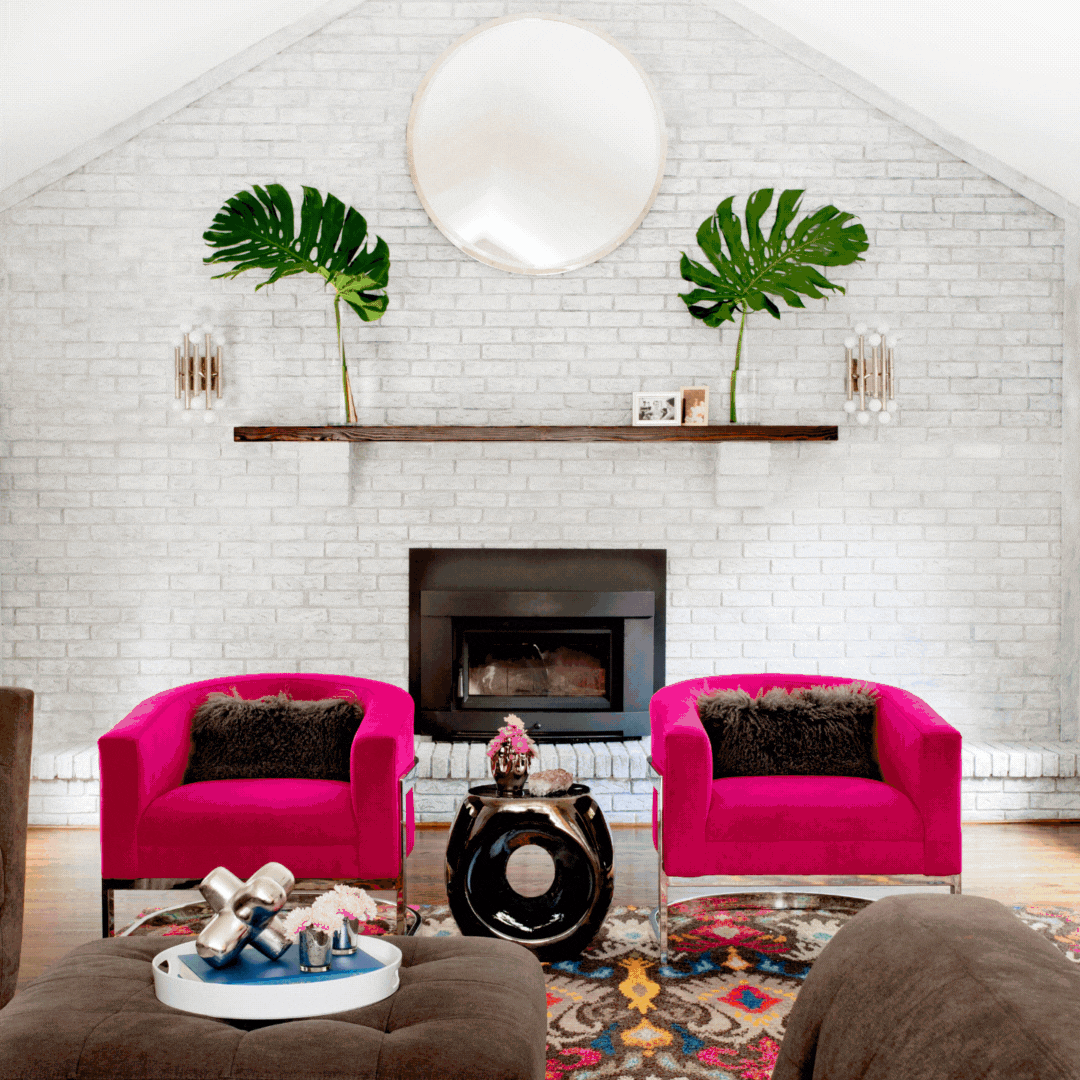 Living room with a white brick wall, two pink armchairs with dark pillows, a round black side table with a pink flower arrangement, a fireplace, and a decorative rug.