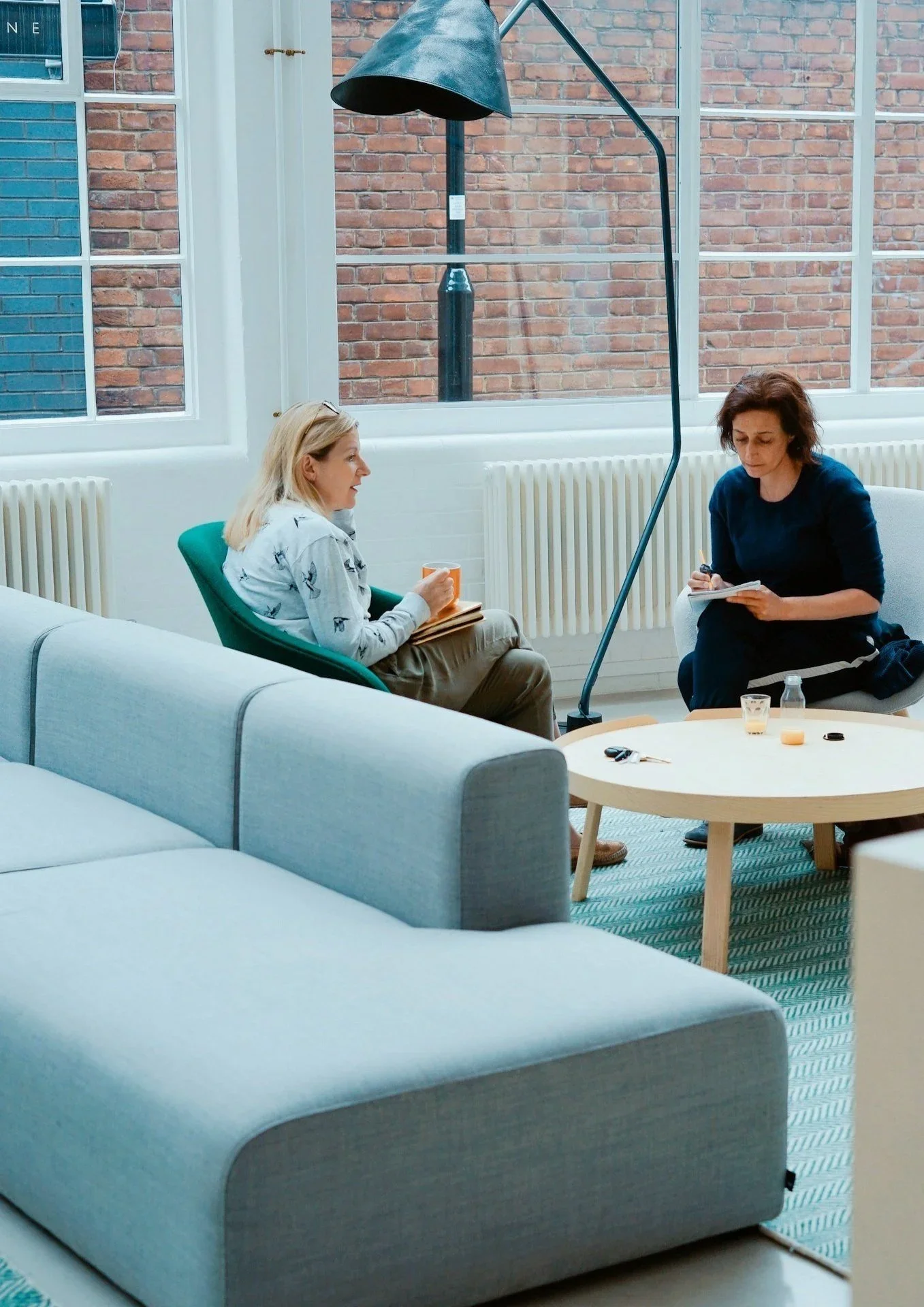 Two women sit in a modern room, having a conversation. One is holding a mug, and the other is taking notes. A large window with a brick wall outside is behind them, and a tall, black floor lamp is nearby.