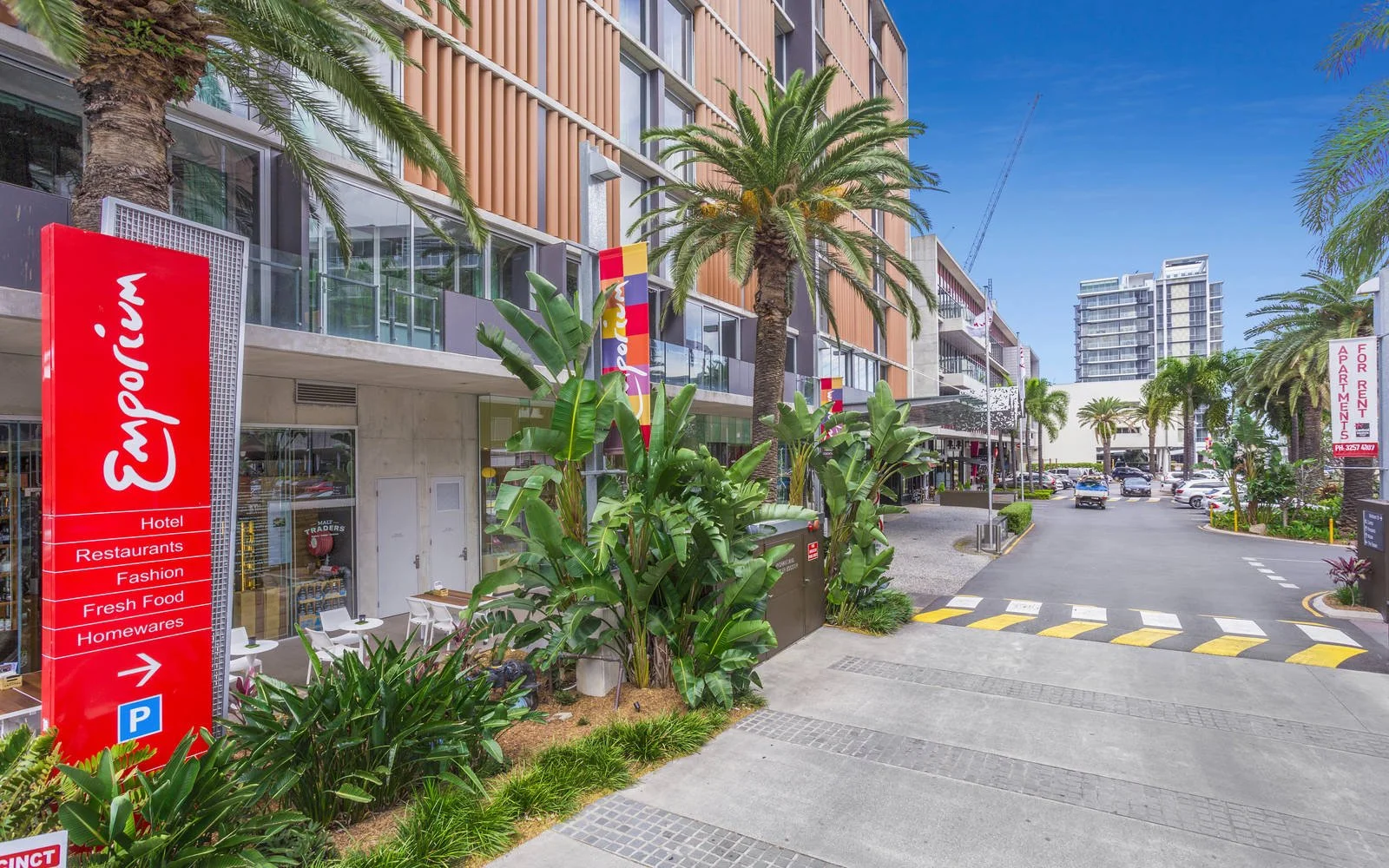 Street view of a modern hotel with a red Emporium sign, lush green palm trees, and tropical plants. Cars are parked along the street, and there are high-rise buildings in the background under a clear blue sky.