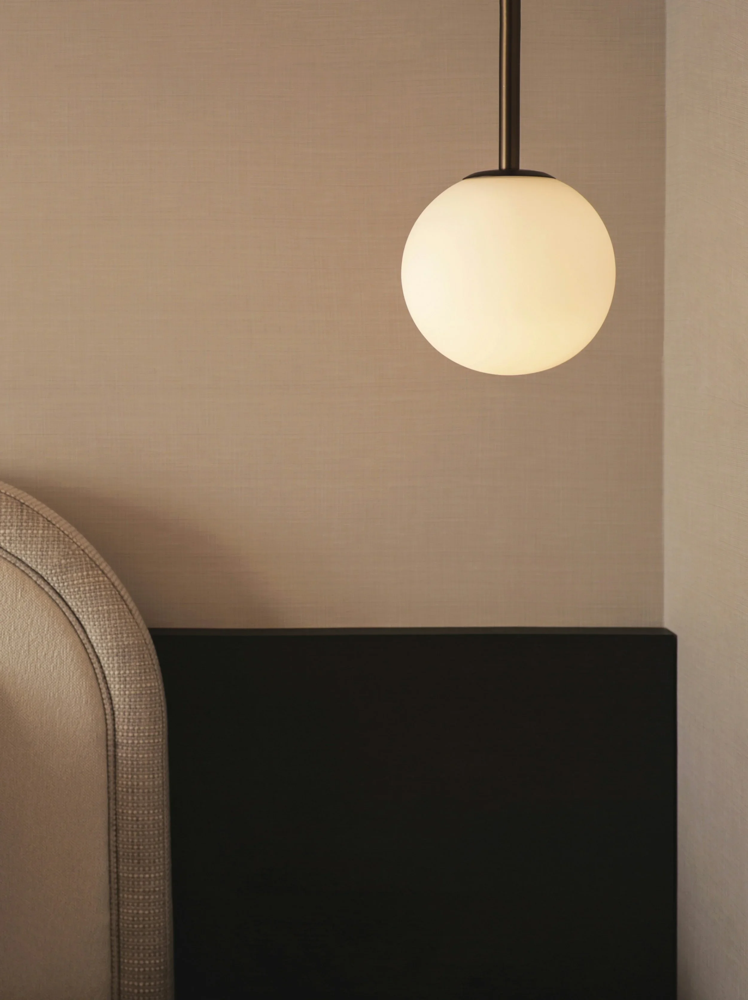 A hanging pendant light with a white, spherical glass shade in a modern interior, next to a beige upholstered headboard and a textured wall.