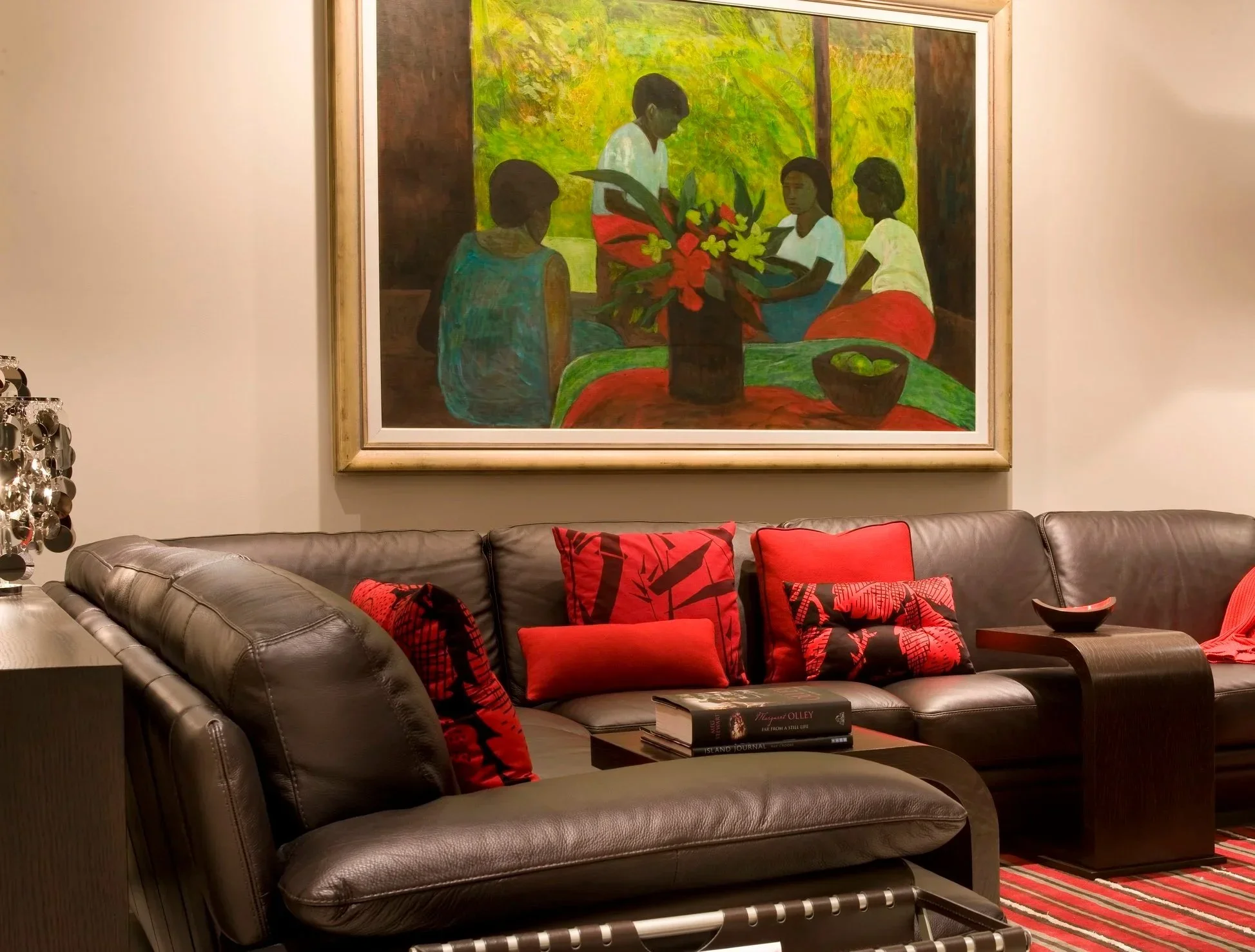 Living room with dark leather sectional sofa decorated with red and black pillows and a glass coffee table with books, in front of a large framed painting of four women gathered around a table with a flower vase, set against a green background.