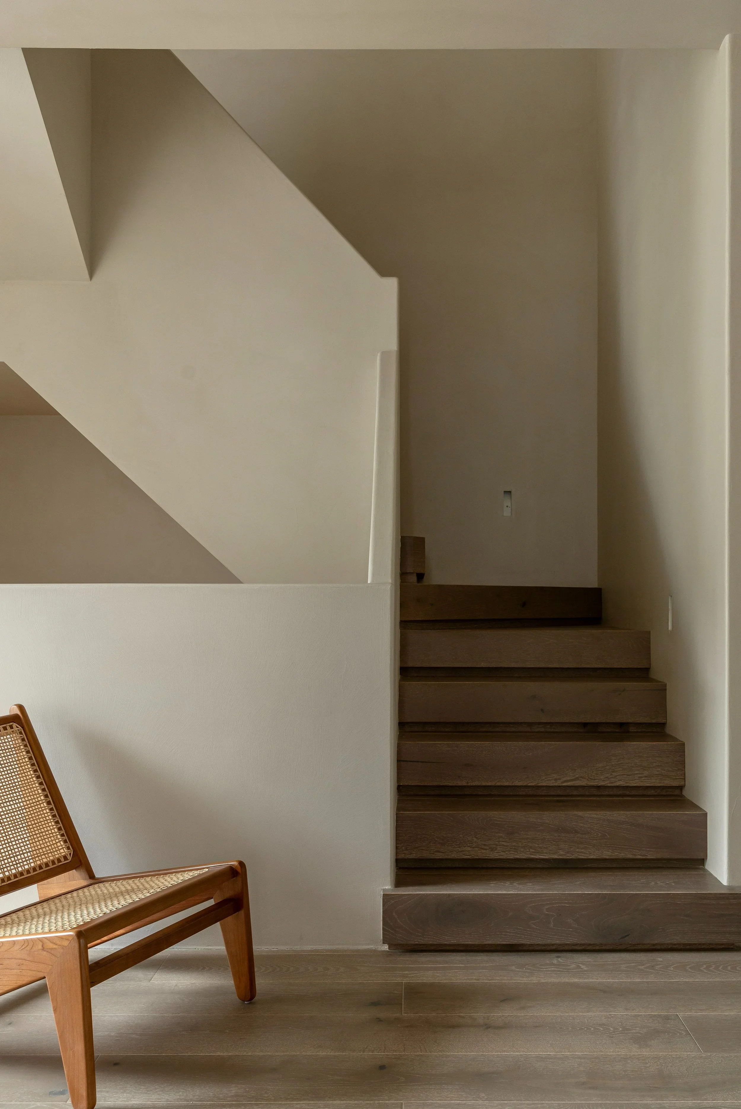 Interior view of a staircase with wooden steps, a partial view of a wooden chair with a woven seat, and plain beige walls in a minimalist style.