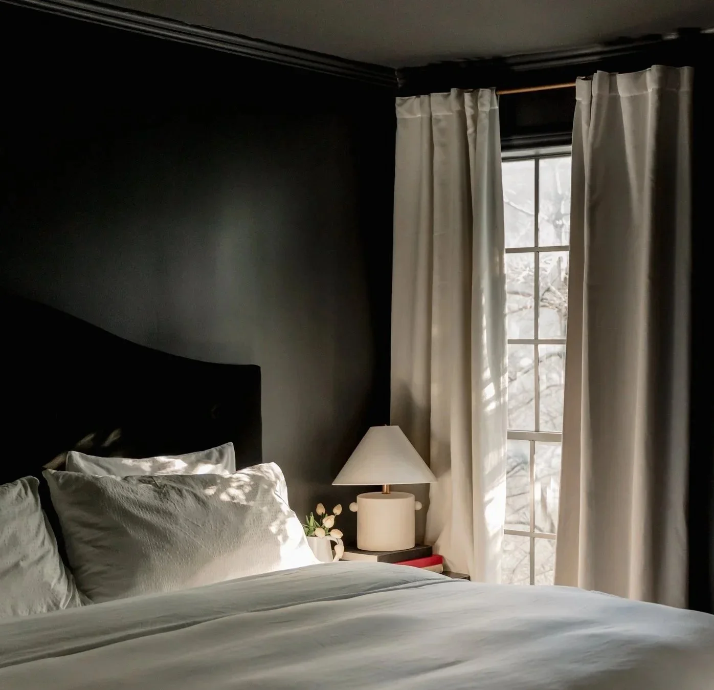 A cozy bedroom with a dark accent wall, a bed with white pillows and sheets, a bedside table with a lamp, and a window with white curtains letting in natural light.