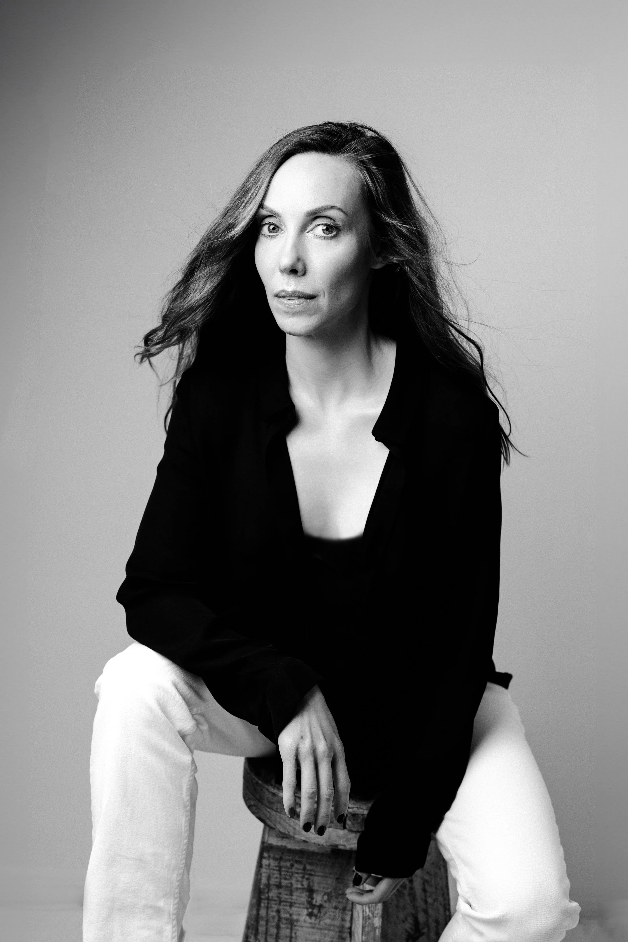 Black and white portrait of a woman with long wavy hair, sitting on a wooden stool, wearing a black top and light-colored pants, against a plain background.