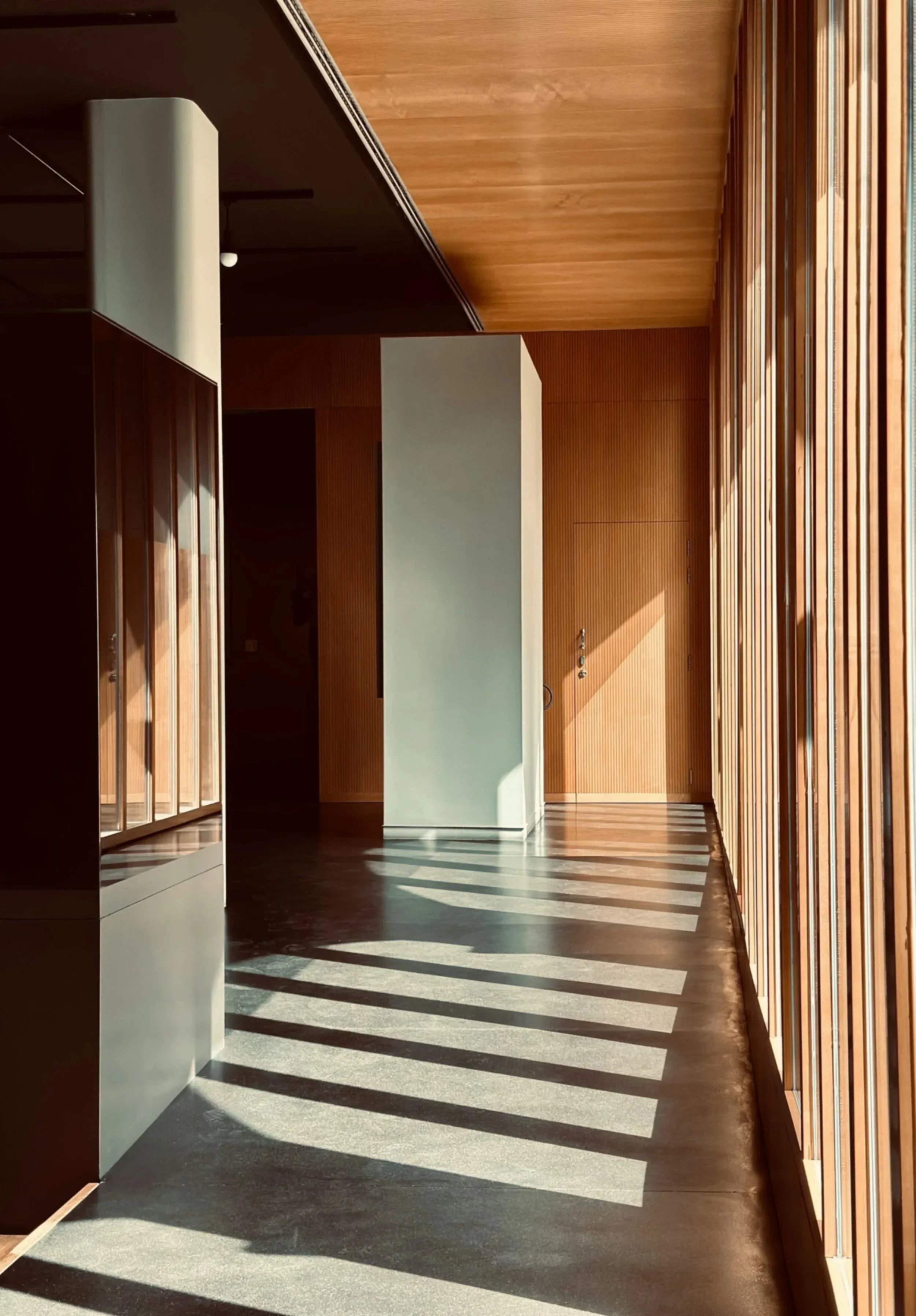 Interior hallway with wooden wall panels and strips of sunlight creating shadows on the concrete floor.