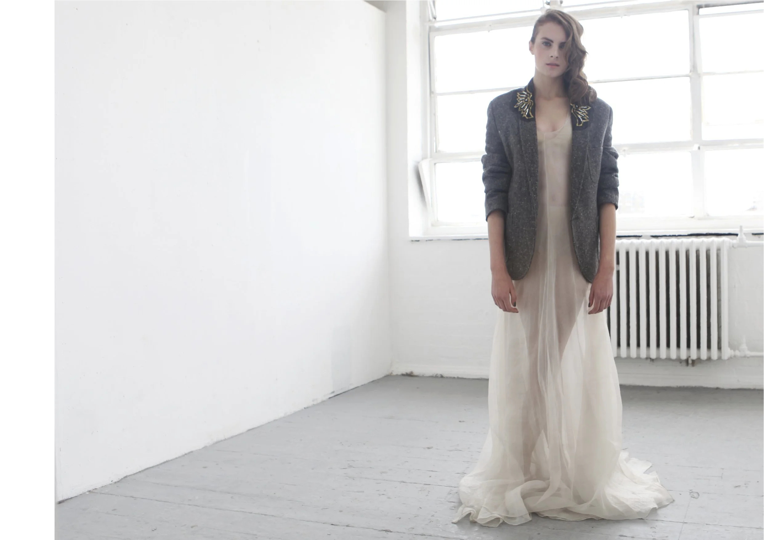 A woman standing in a bright, white room with a large window and radiator, wearing a beige dress and a dark blazer with decorative embellishments on the shoulders.