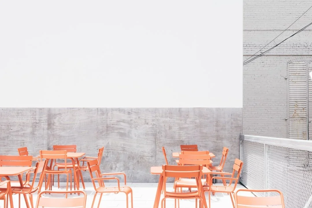 Outdoor terrace with orange chairs and tables against a gray concrete wall and a white brick building wall in the background.