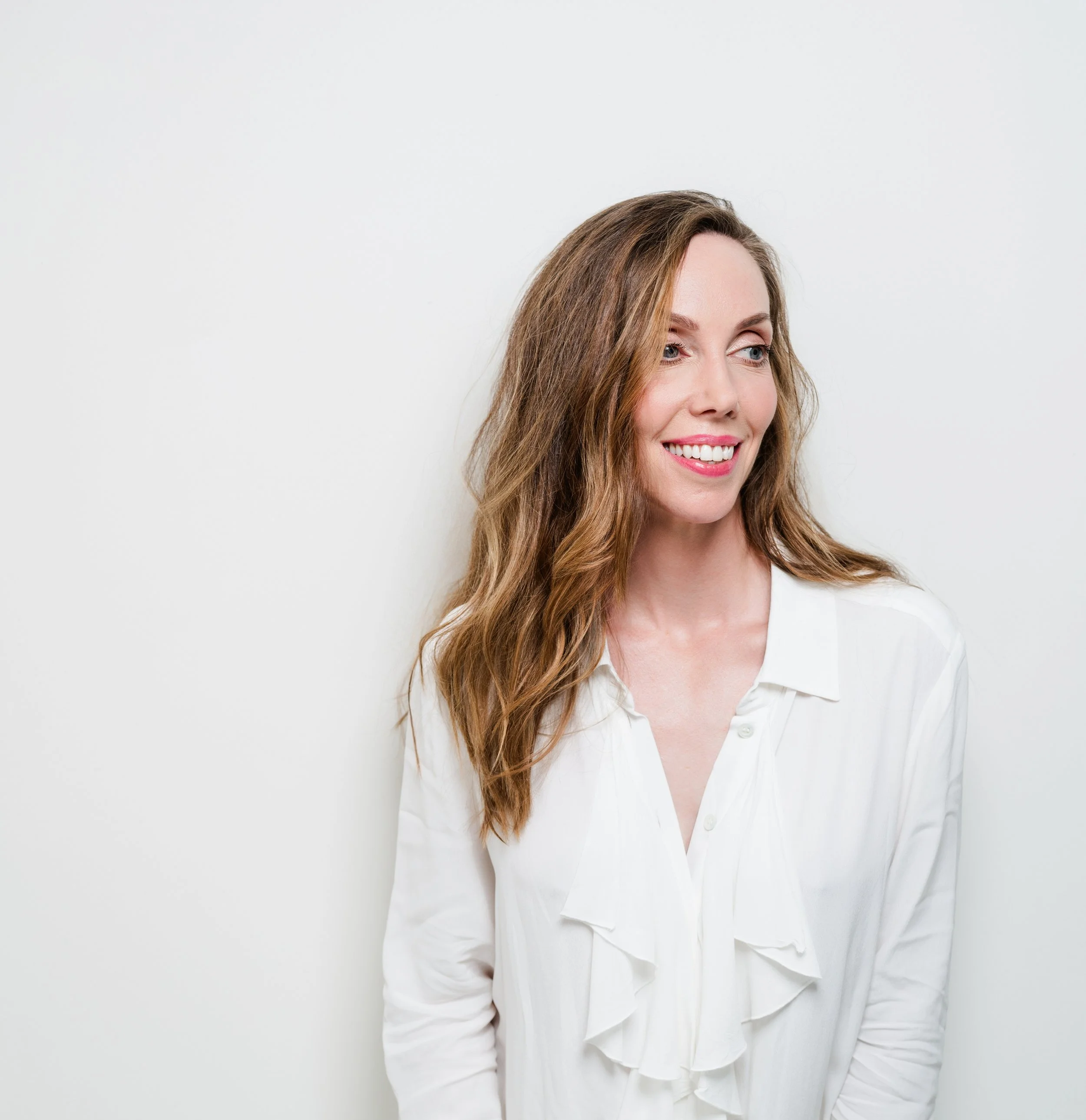 A woman with long wavy brown hair smiling, wearing a white blouse, standing against a plain white background.