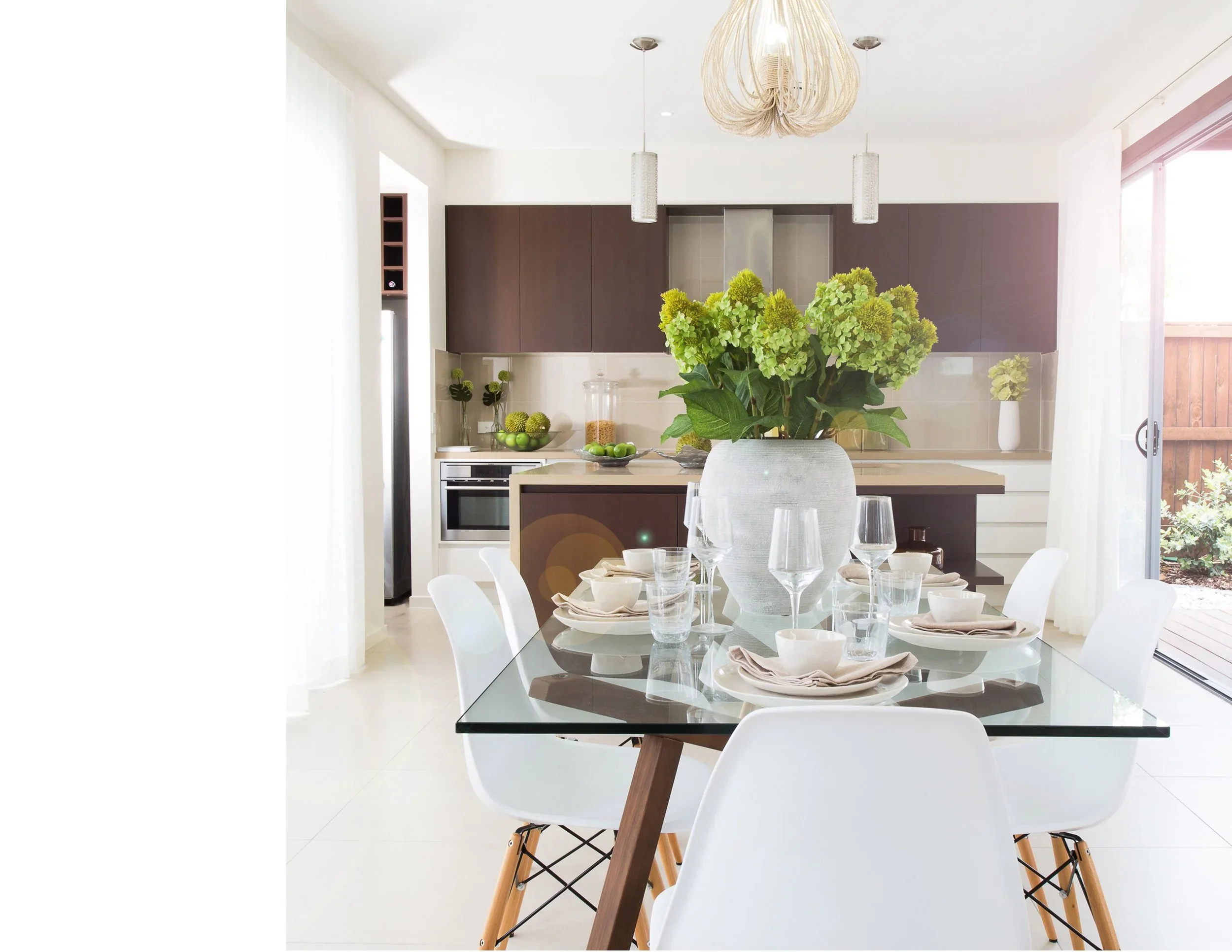 A modern kitchen and dining area with a glass table set for four, featuring white dishes, glasses, and beige napkins. A large white vase with green hydrangea flowers is at the center of the table. The kitchen in the background has dark brown cabinets