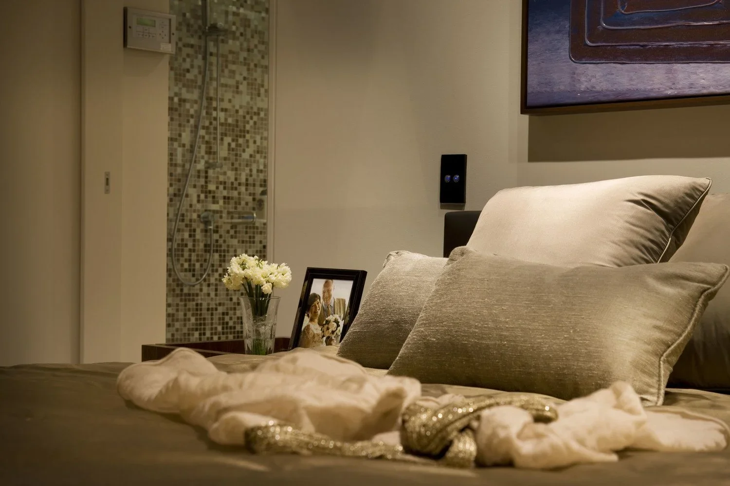 Close-up of a cozy bed with beige pillows in a hotel room, with a framed wedding photo, a vase of white flowers, and a patterned blanket draped over the bed. In the background, there is a wall with a mounted TV and a bathroom with a shower visible.