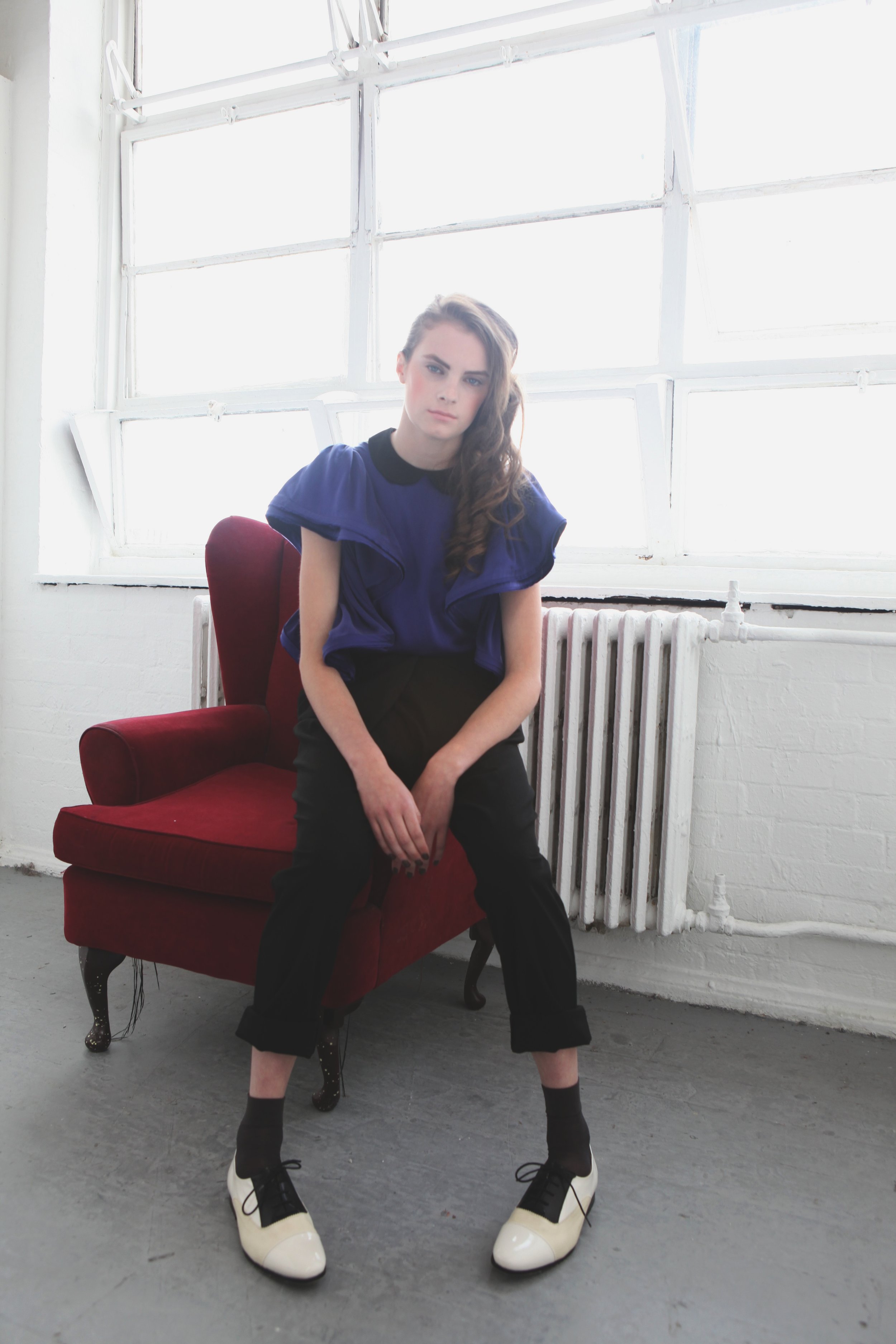 A young woman with long, wavy brown hair sitting on a red vintage armchair in a room with white brick walls and large industrial windows. She wears a royal blue top, black pants, black socks, and two-tone white and black shoes.