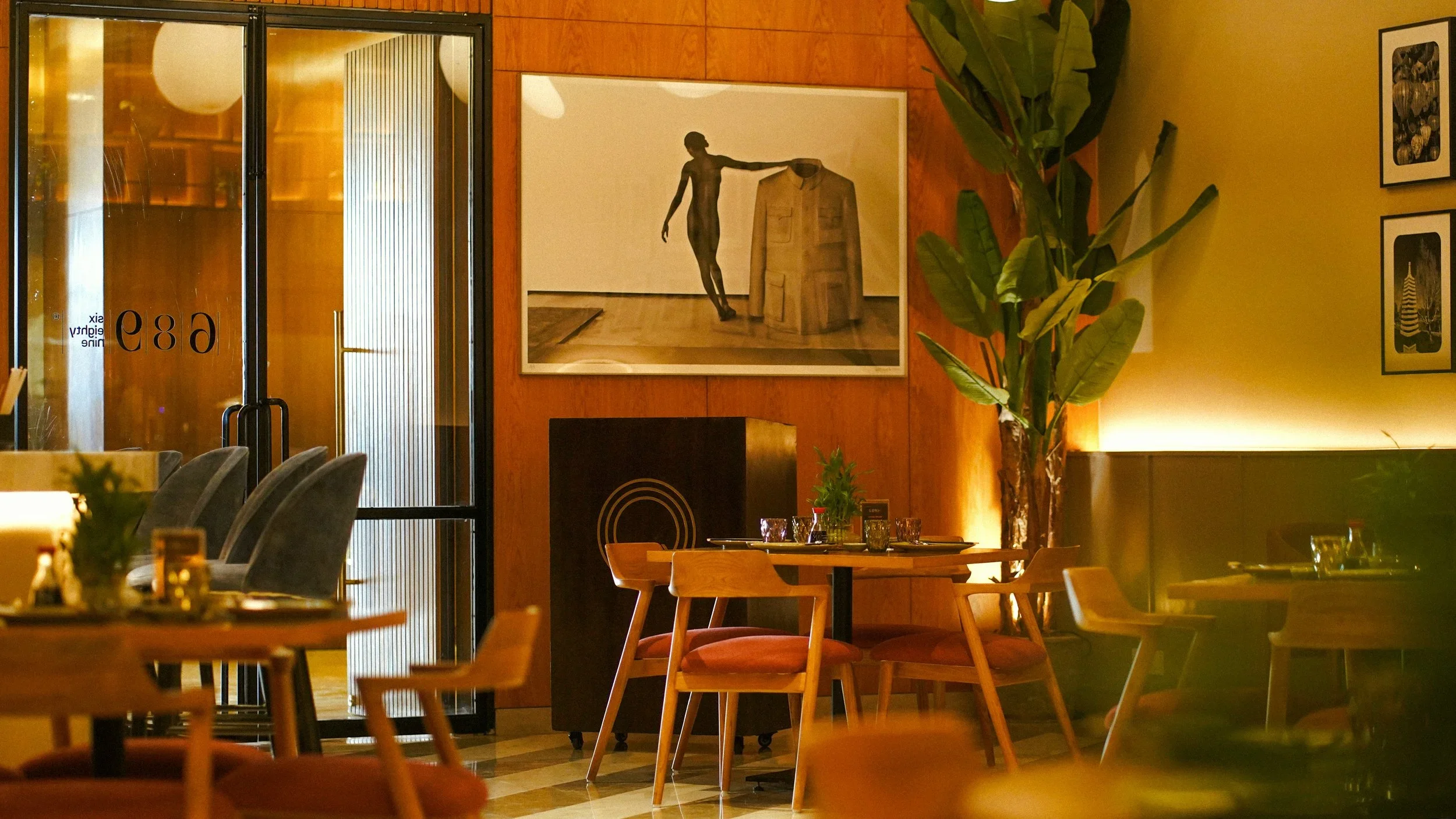 Interior of a restaurant with wooden furniture, a large plant, wall art, and warm lighting.