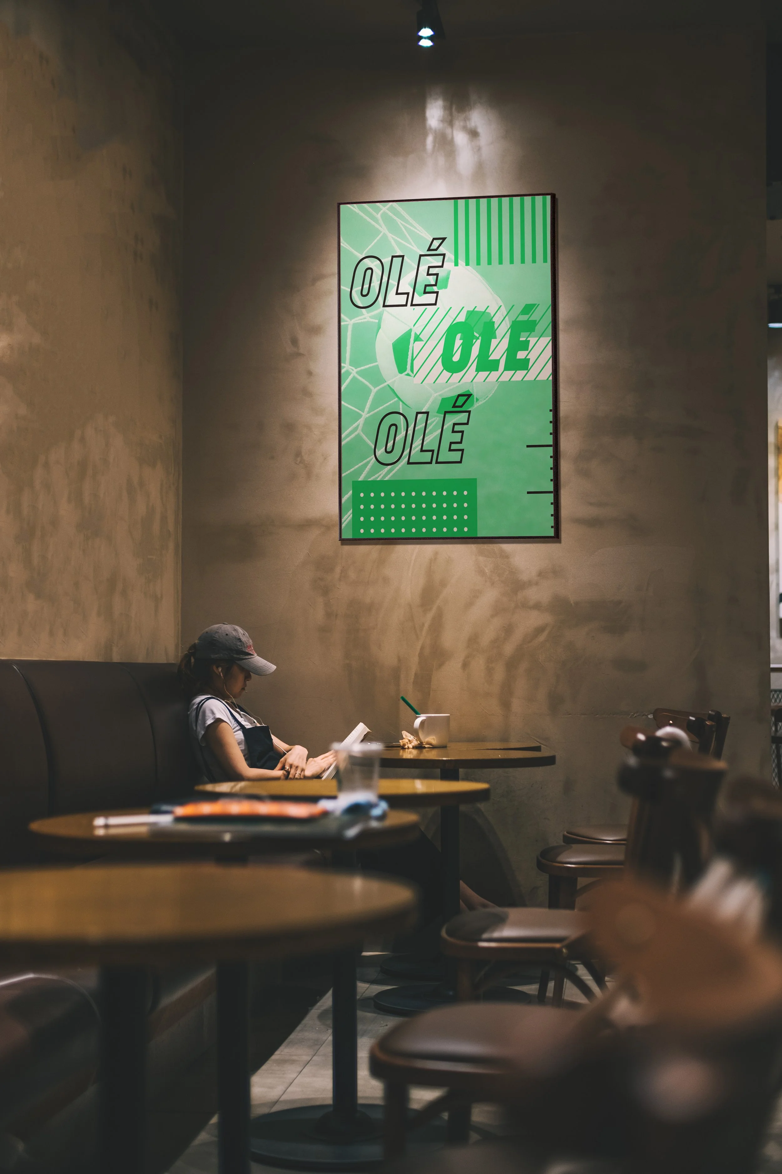 A young girl sitting alone at a table in a cafe, reading a book. The cafe has beige walls, wooden tables, and chairs. There is a large green and white abstract poster on the wall that says 'OLÉ' repeated three times.
