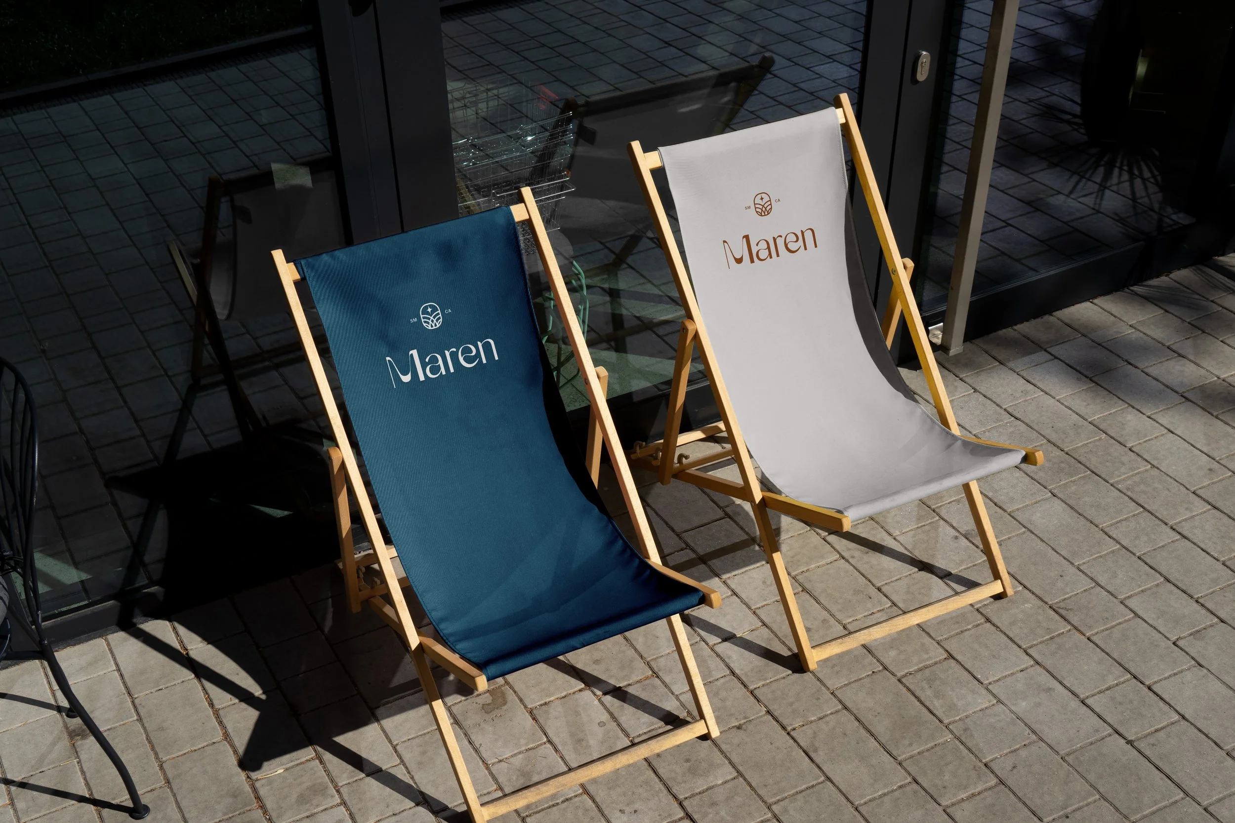 Two wooden deck chairs with fabric seats, one blue and one white, with the word "Maren" written on each, are placed on a brick patio outside a building with large glass doors.