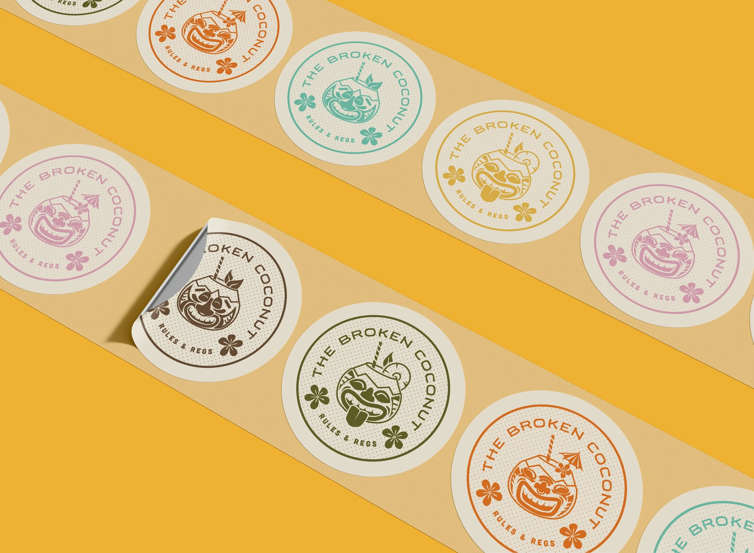 Sheet of colorful round stickers featuring carved coconut faces and the text ‘The Broken Coconut Rules & Regs’ in various colors. One sticker is partially peeled.