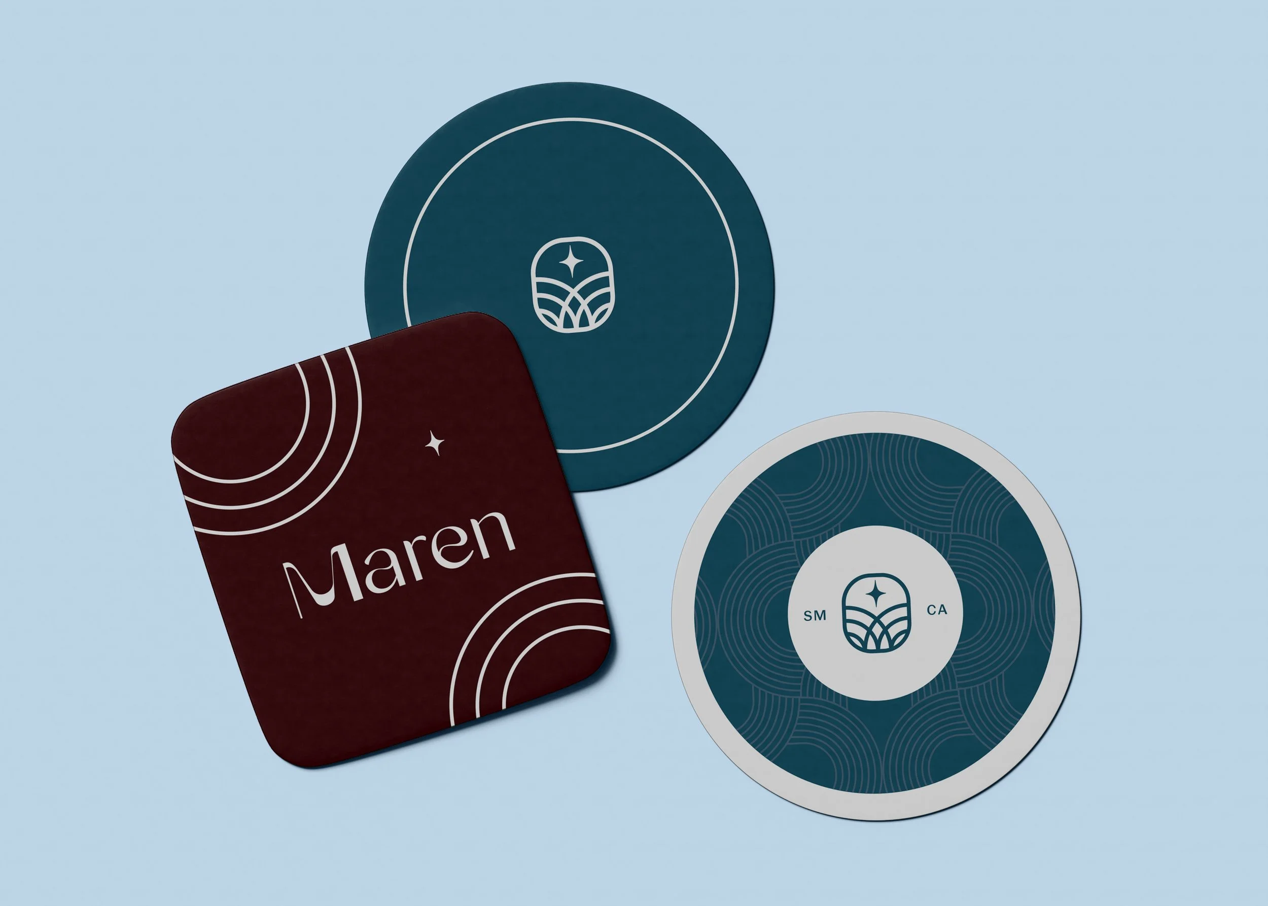 Three round coasters with blue and white designs, and a square dark red coaster with white text reading ‘Maren’ and white curved line patterns.