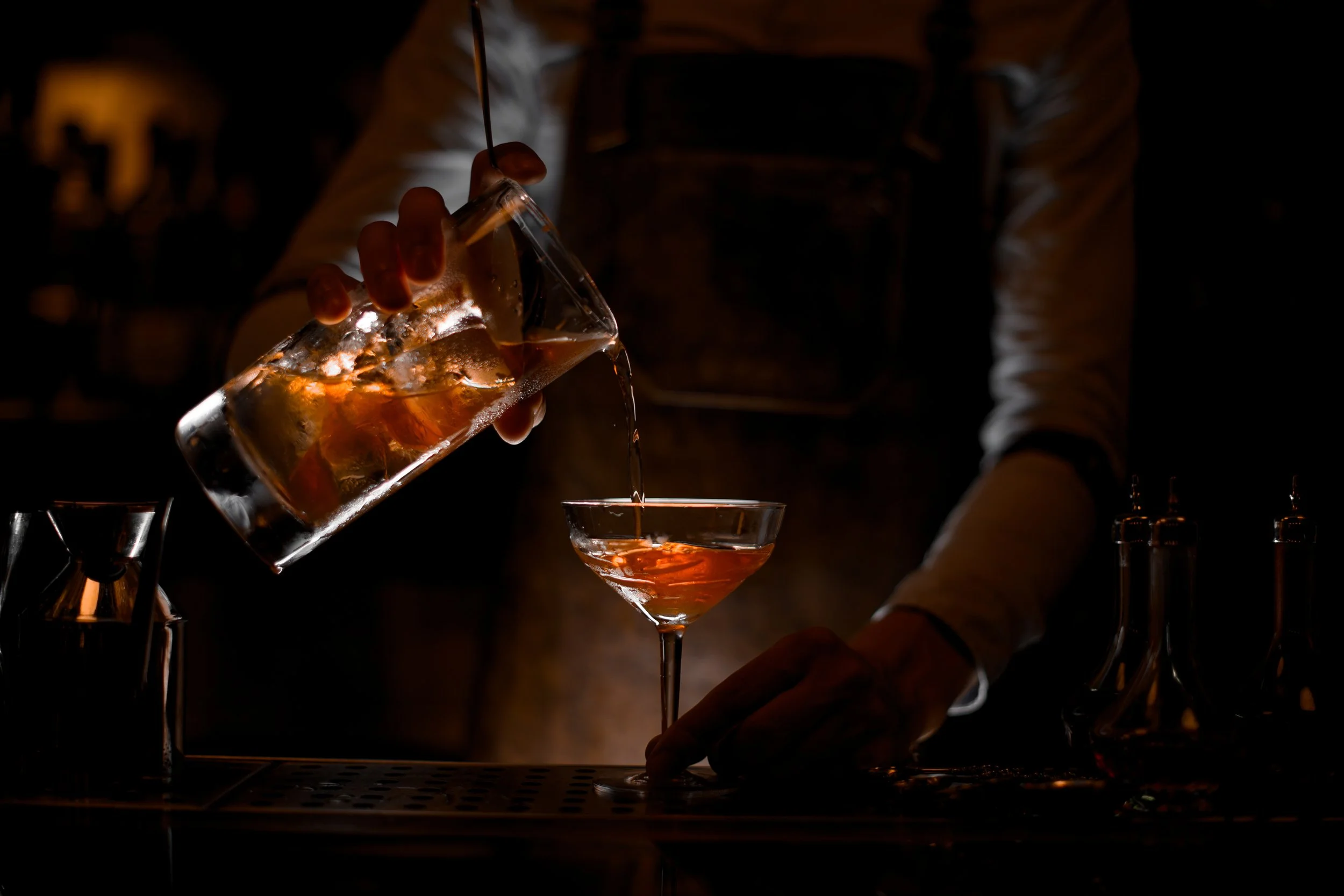 A bartender pouring a drink from a glass pitcher into a cocktail glass at a dimly lit bar.
