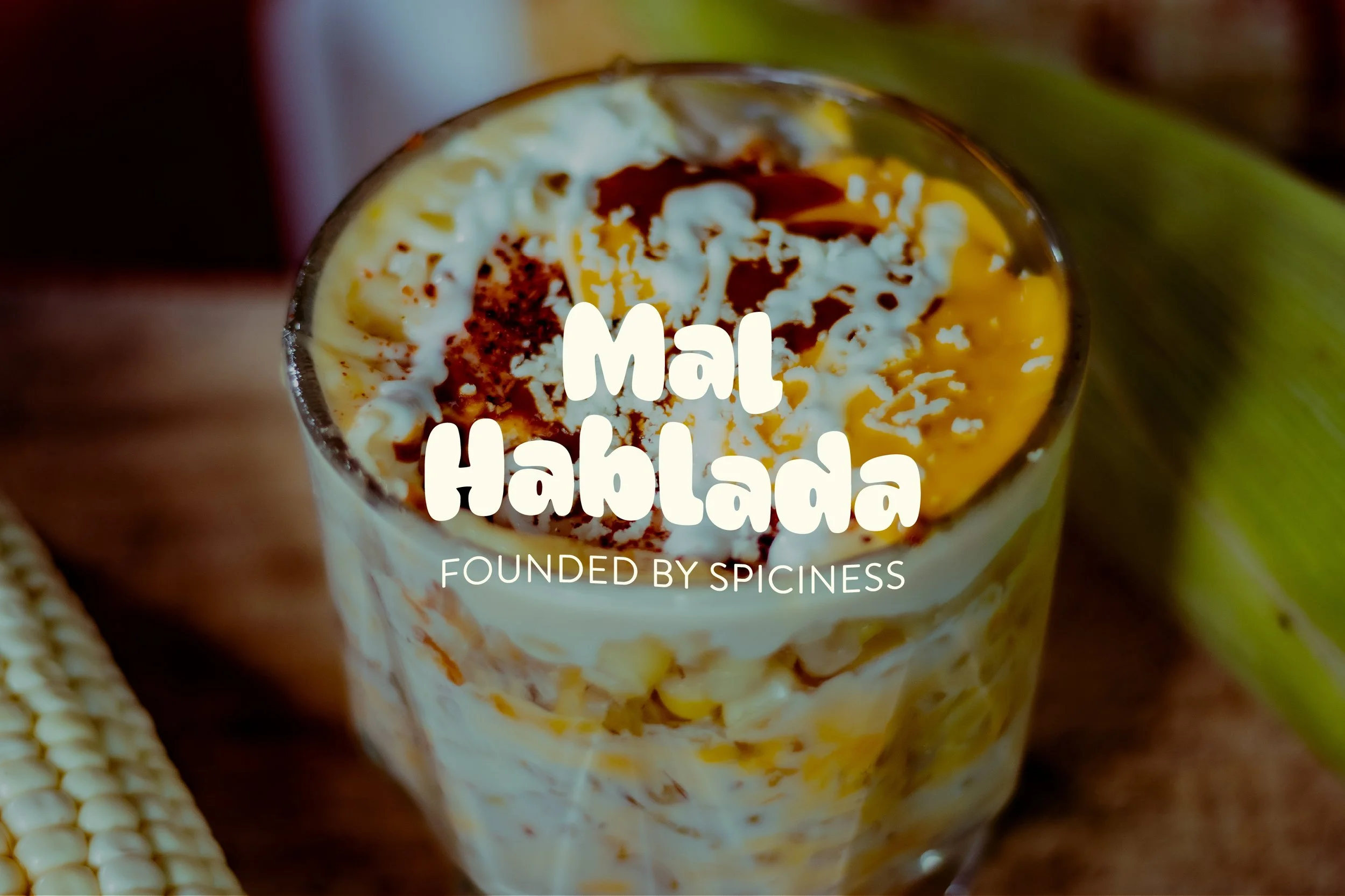 Close-up of a glass filled with esquite (Mexican street corn), with a green plant and corn on the cob in the background. Overlaid text reads ‘Mal Hablanda’ and ‘Founded by Spiciness.