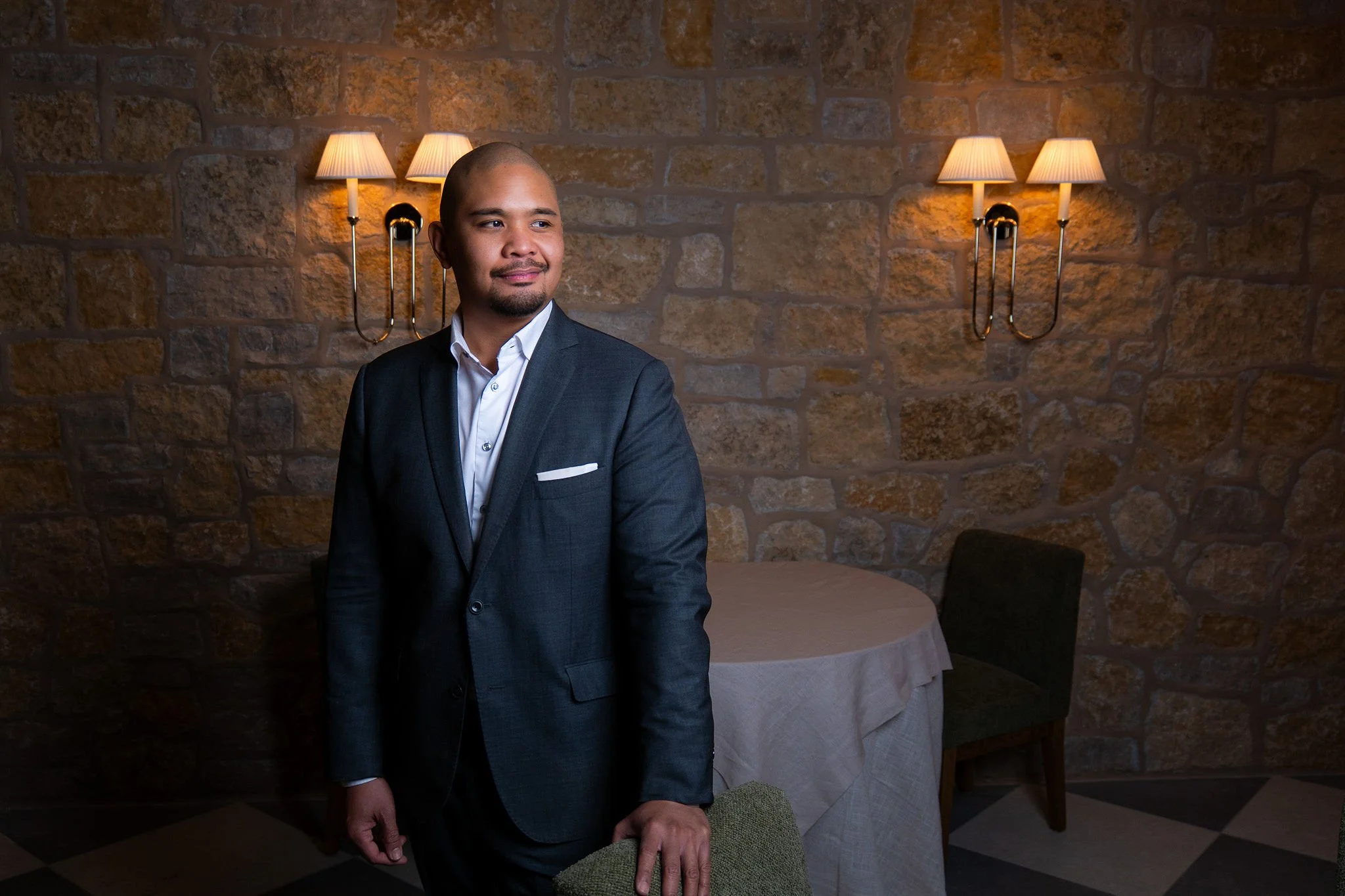 A man in a dark suit and white shirt standing in a room with stone walls and wall-mounted lamps, with a round table covered with a cloth and chairs in the background.