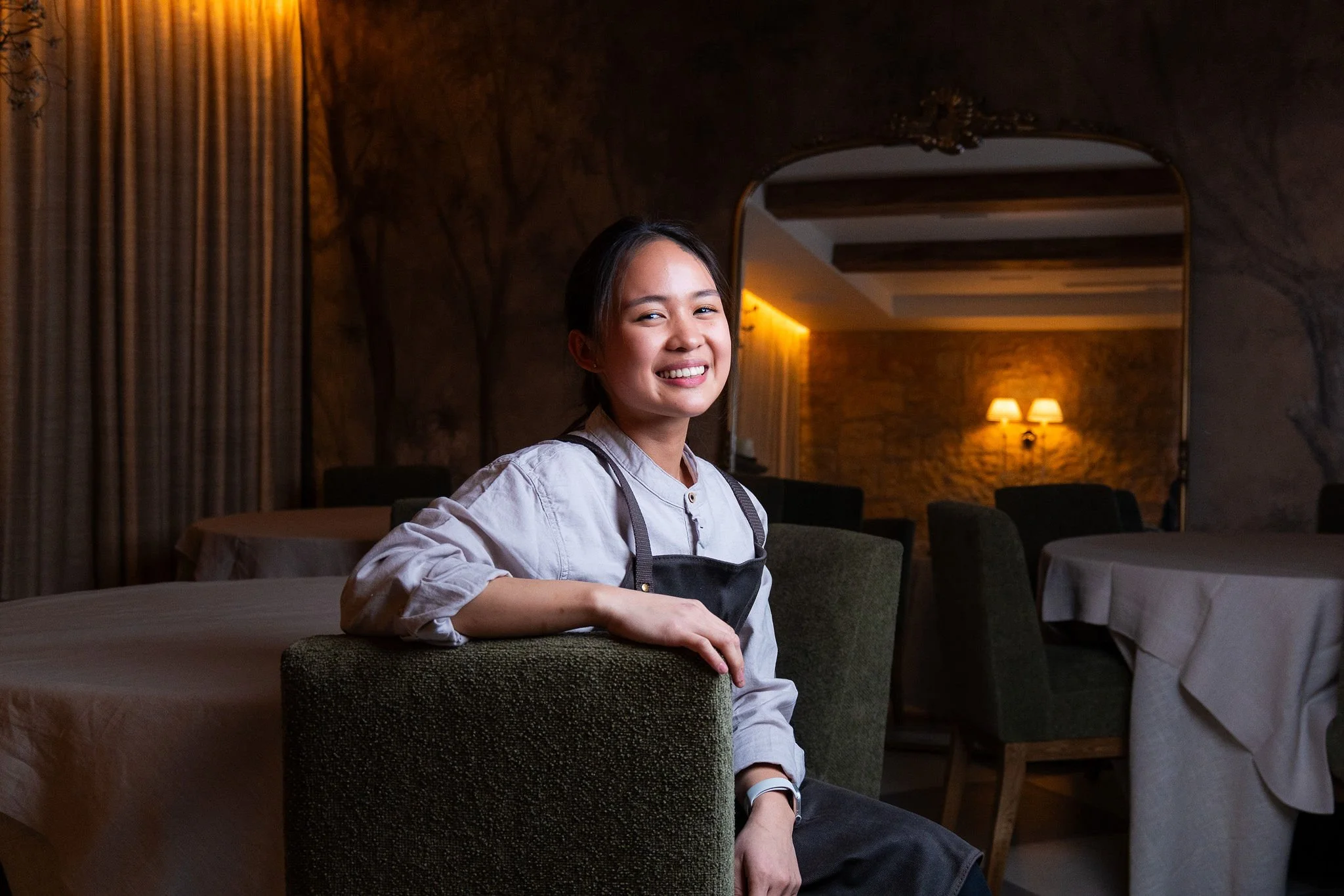 A smiling woman sitting at a table in a warmly lit restaurant or cafe with dark decor and stone walls.