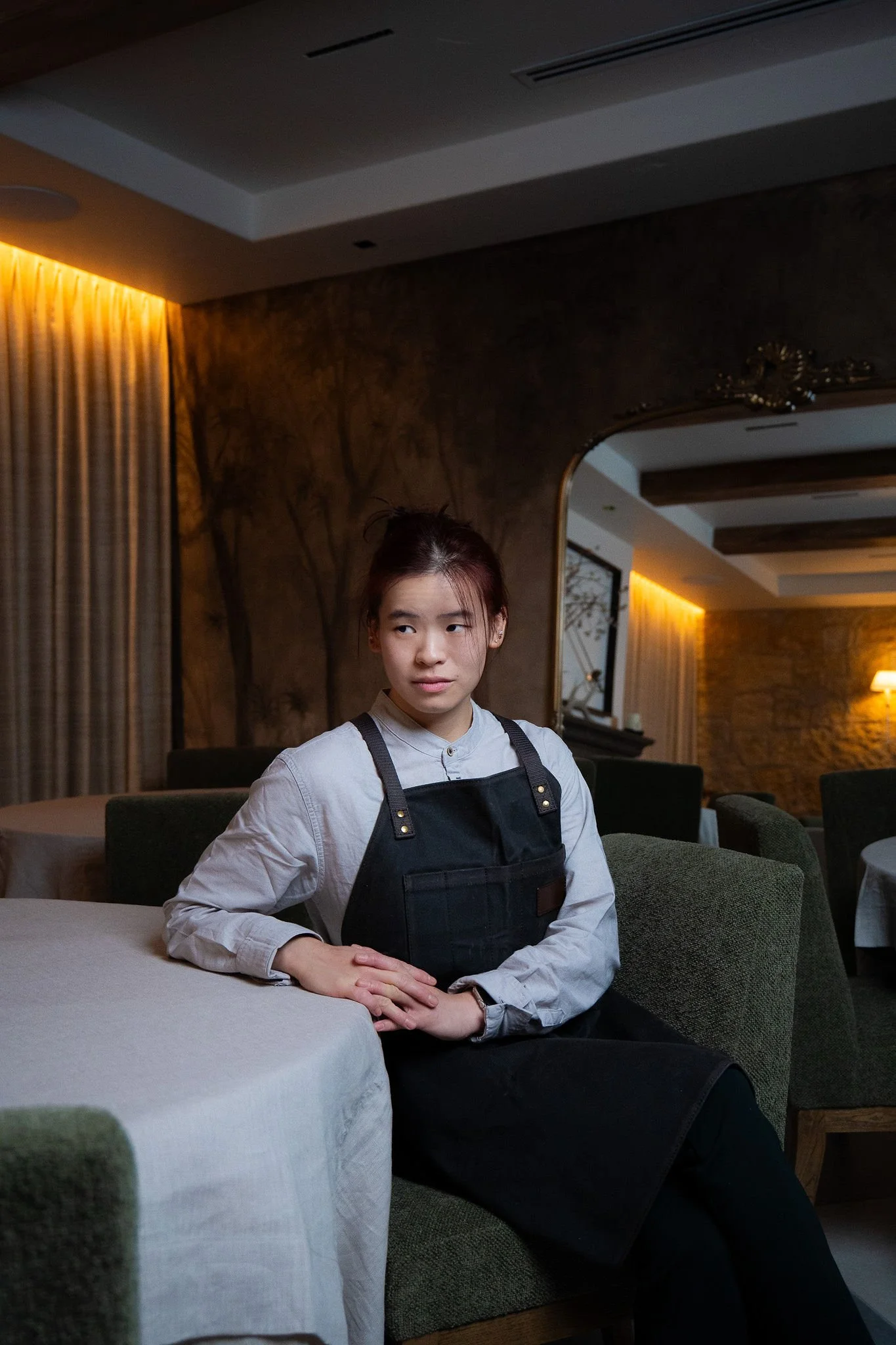A young woman sitting at a table in a restaurant or cafe, wearing a white shirt and black apron, with her hands folded on the table, looking to the side.