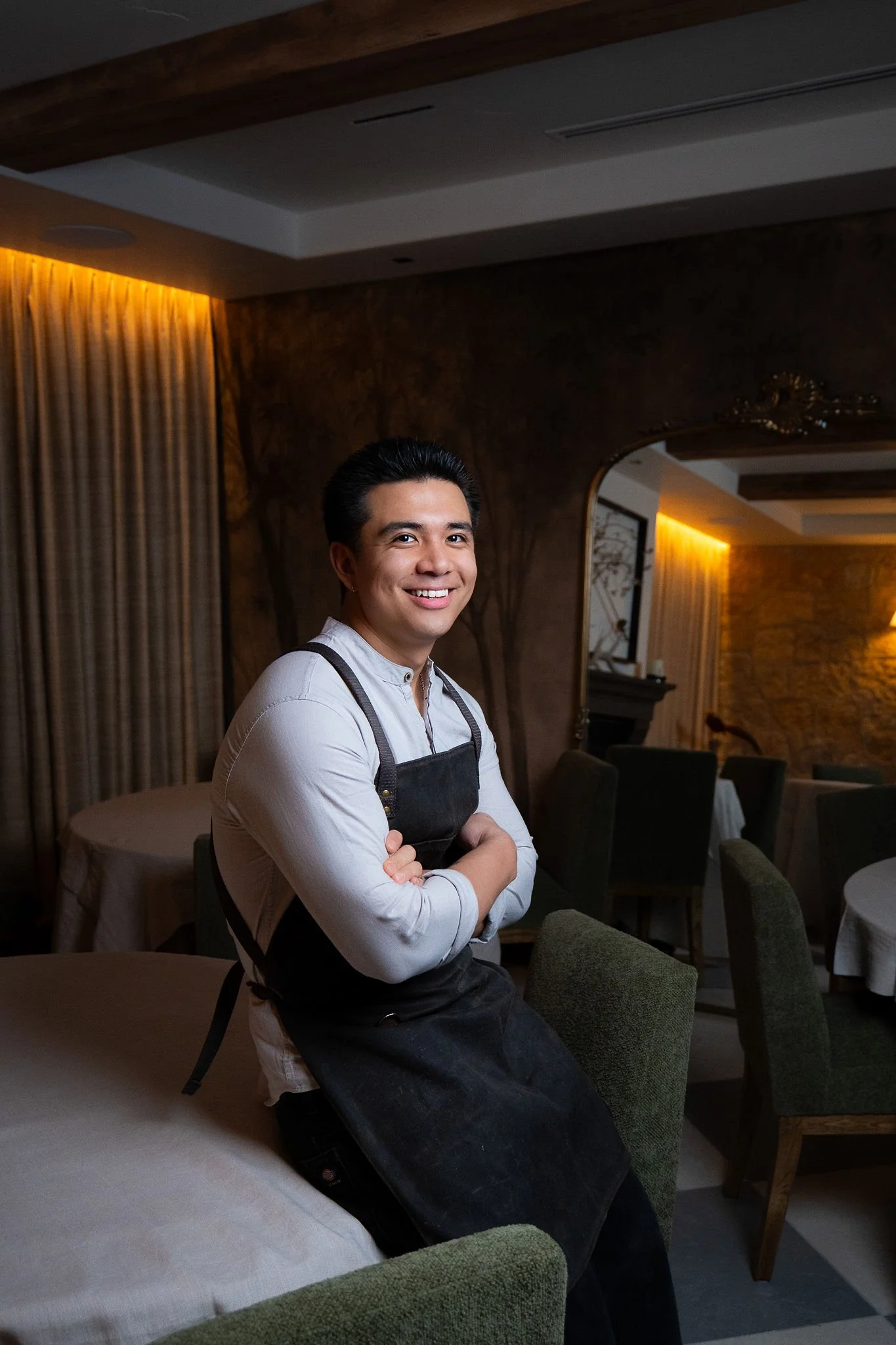 A smiling man in a white shirt and black apron sitting in a restaurant with a warm, softly lit interior.