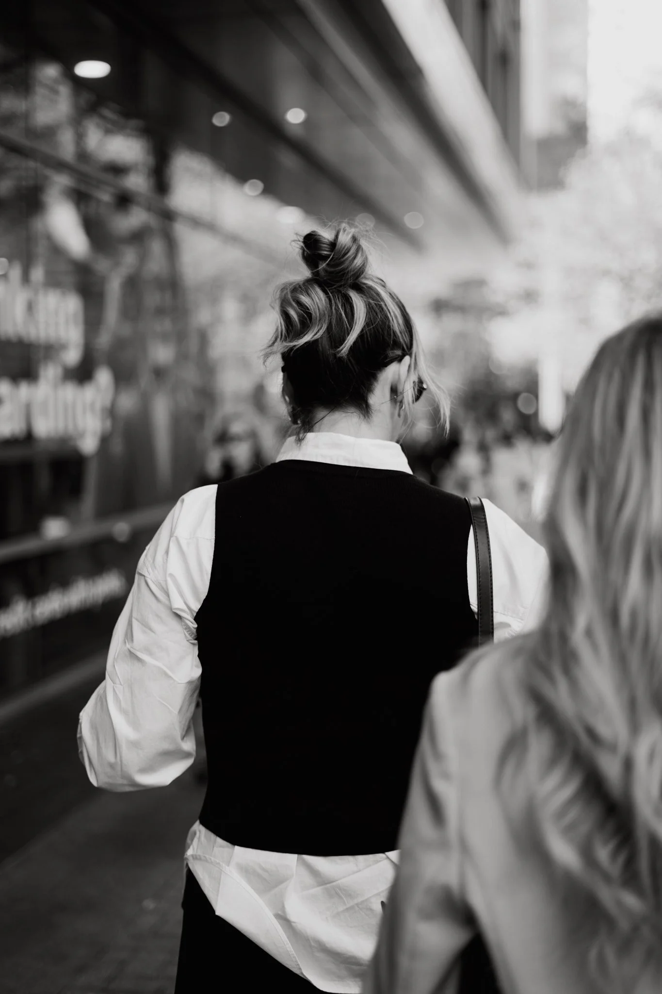 A woman with a top knot hairstyle, wearing a white shirt and a black vest, walking in an urban environment. The photo is in black and white.
