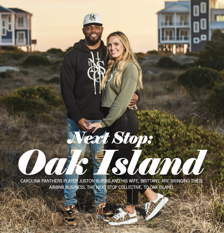 A smiling couple stands close together outdoors on grassy terrain with bushes, near beach houses in the background, during sunset.