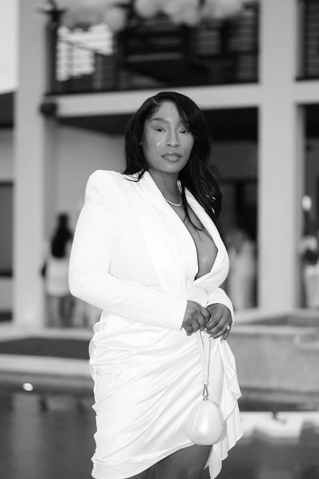 A woman in a white dress holding a small bag, standing outdoors in front of a building.
