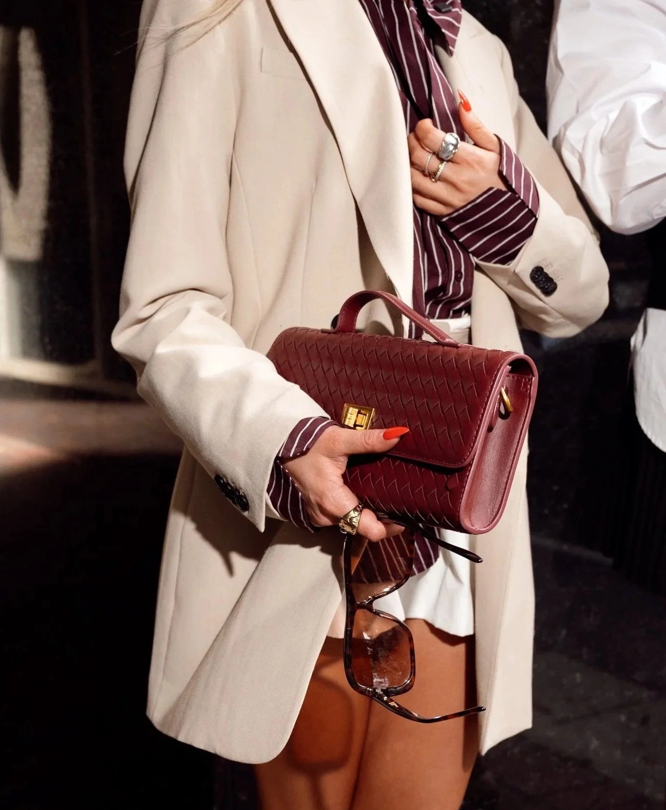 Woman holding a red handbag with sunglasses hanging from her hand, wearing rings, a striped shirt, beige blazer, and brown pants.