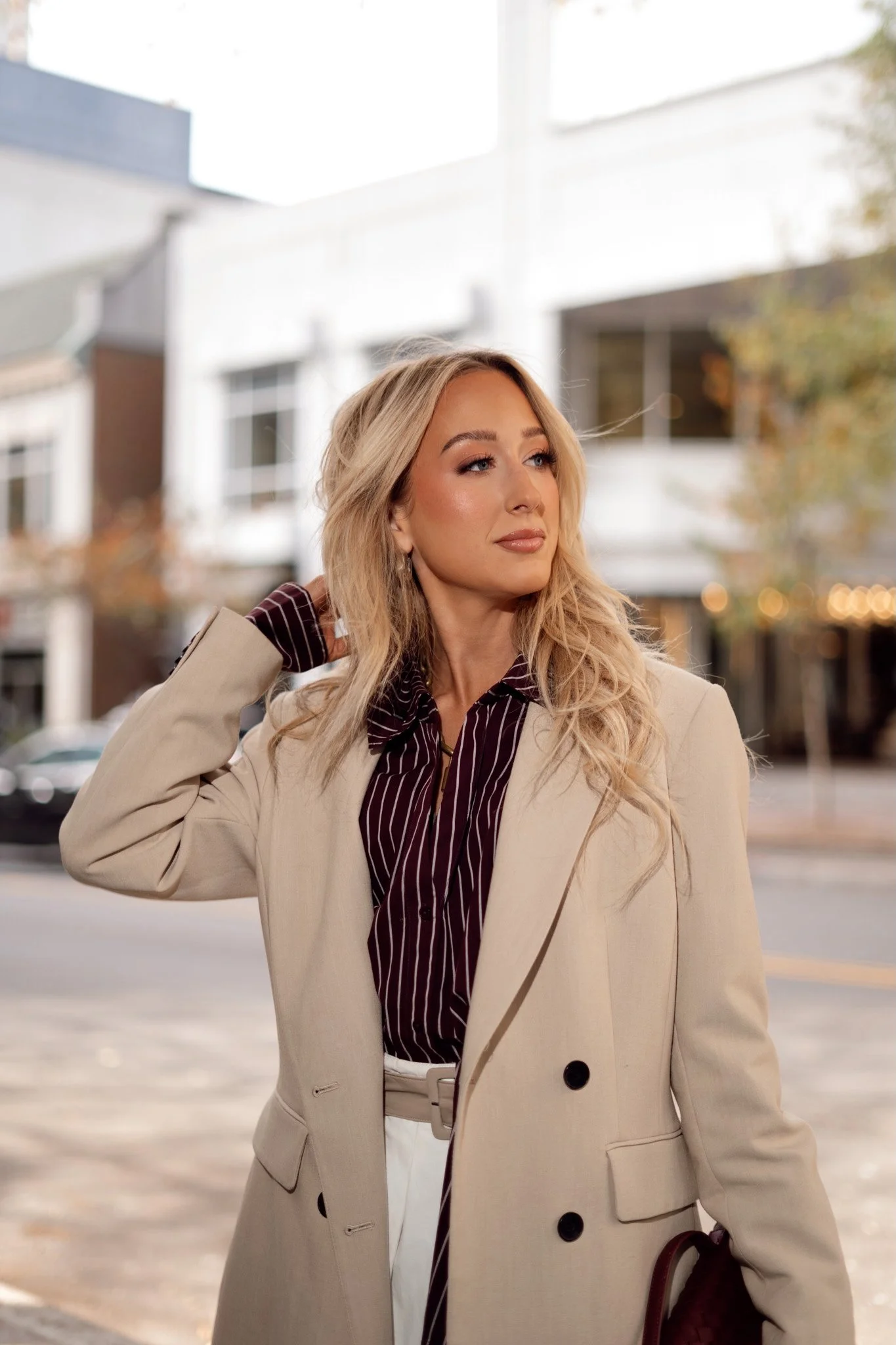 Hannah, a woman with long blonde hair dressed in a beige coat and a dark striped shirt standing outdoors in an urban setting with modern buildings in the background.
