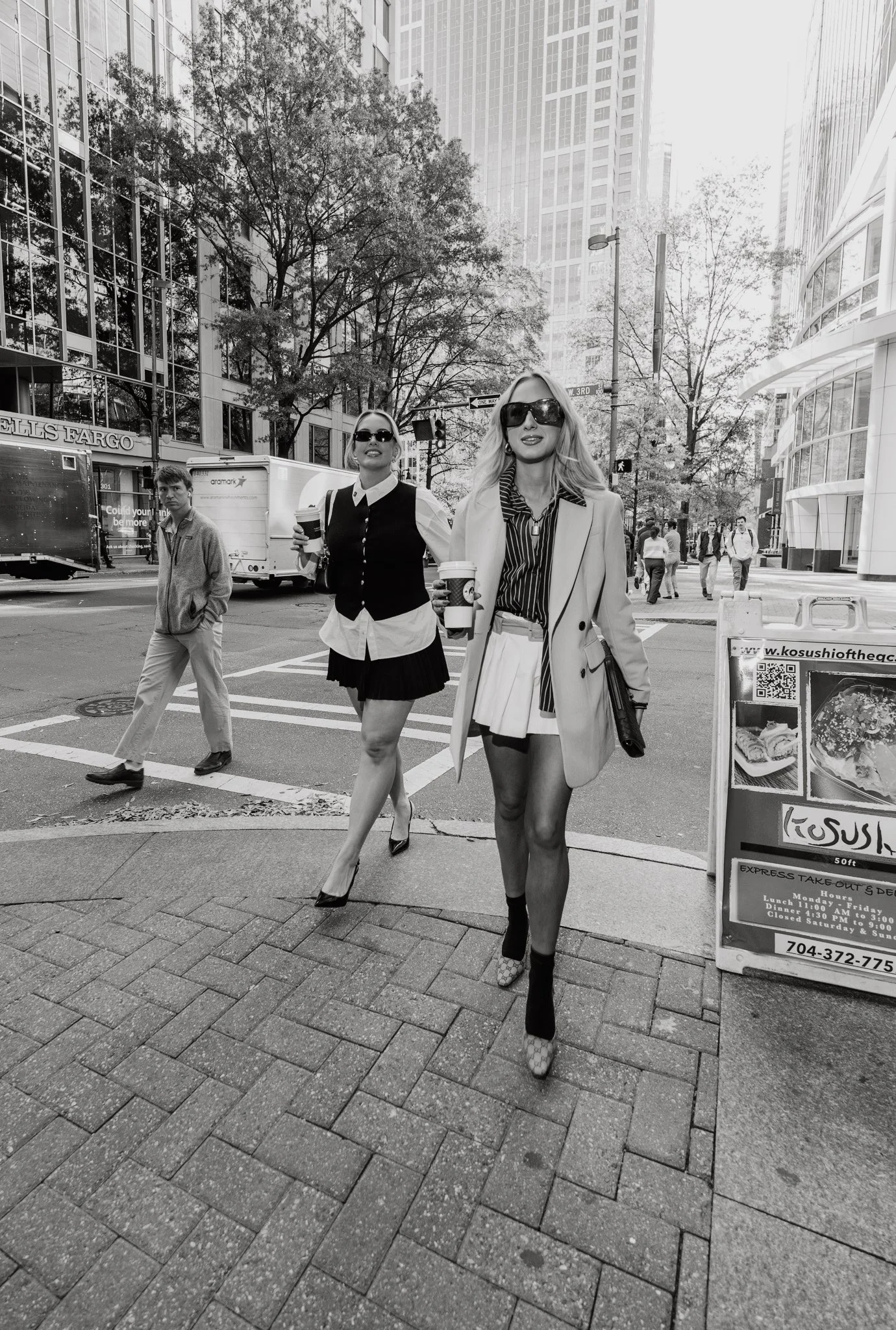 Two fashionable women walking on a city street holding coffee cups, with a man in casual clothes crossing behind them, surrounded by tall buildings and an outdoor dining sign.