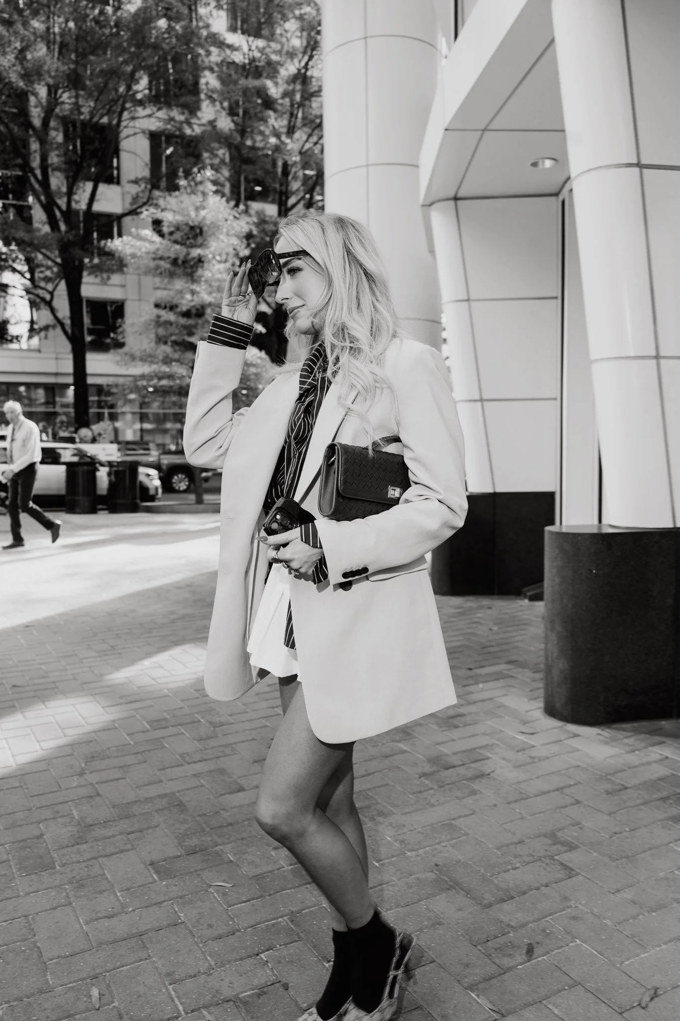 A woman with long wavy hair wearing sunglasses, a light-colored blazer, a striped shirt, and high heels, standing on a city sidewalk holding a small purse and looking to the side.