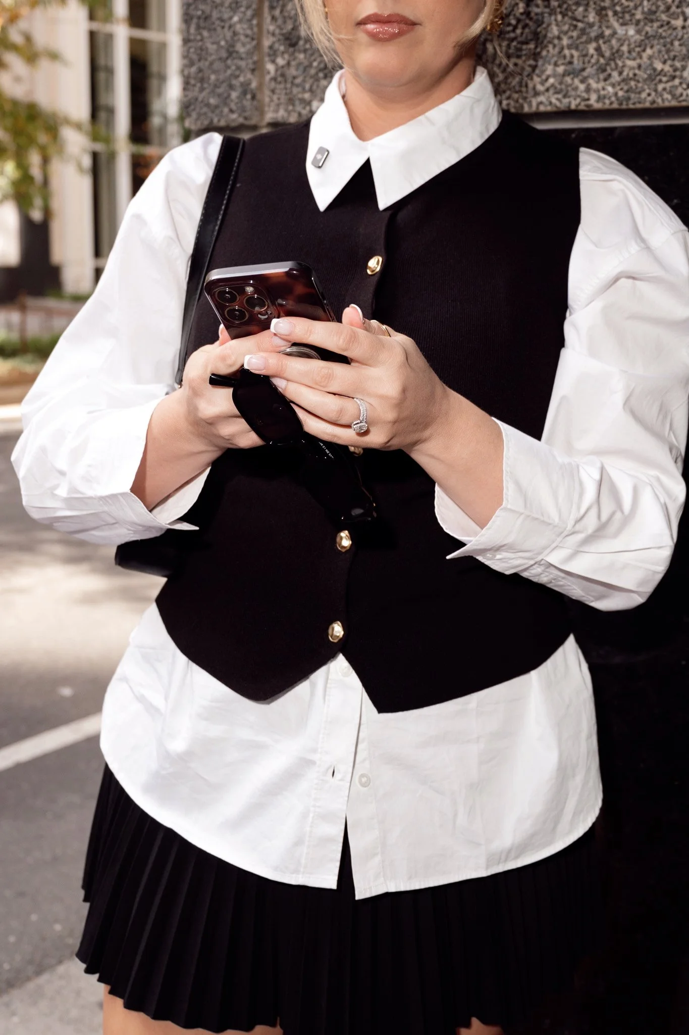 A woman wearing a white blouse, black vest, and black pleated skirt, holding a smartphone with both hands outside near a building.