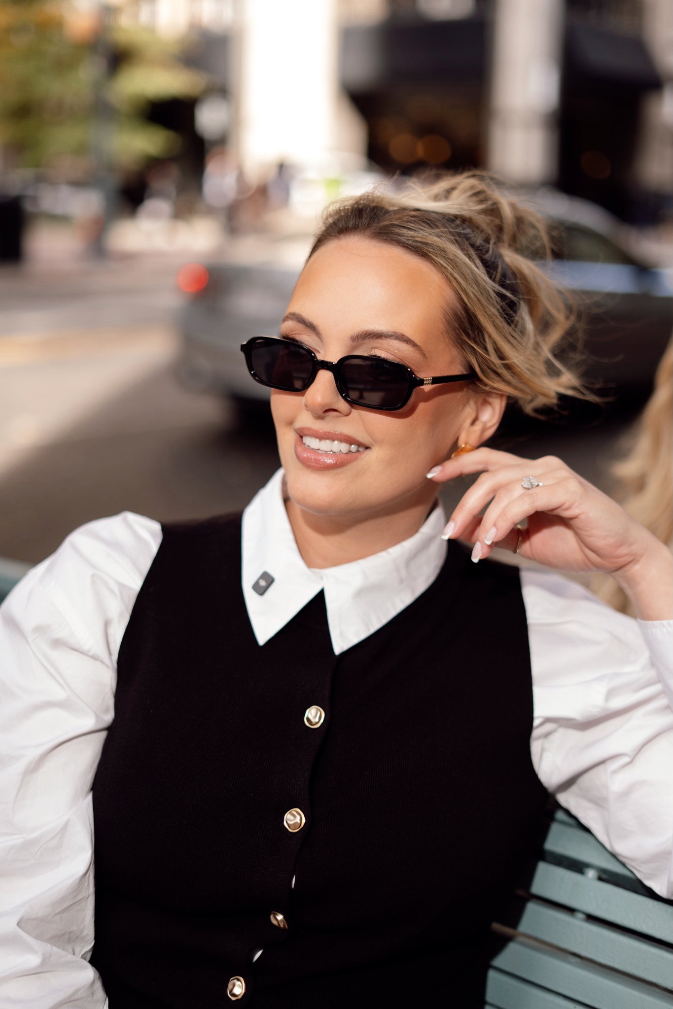 Brittany, a woman with blonde hair, wearing black sunglasses, a white collared shirt under a black vest, is sitting on a bench outdoors, smiling.