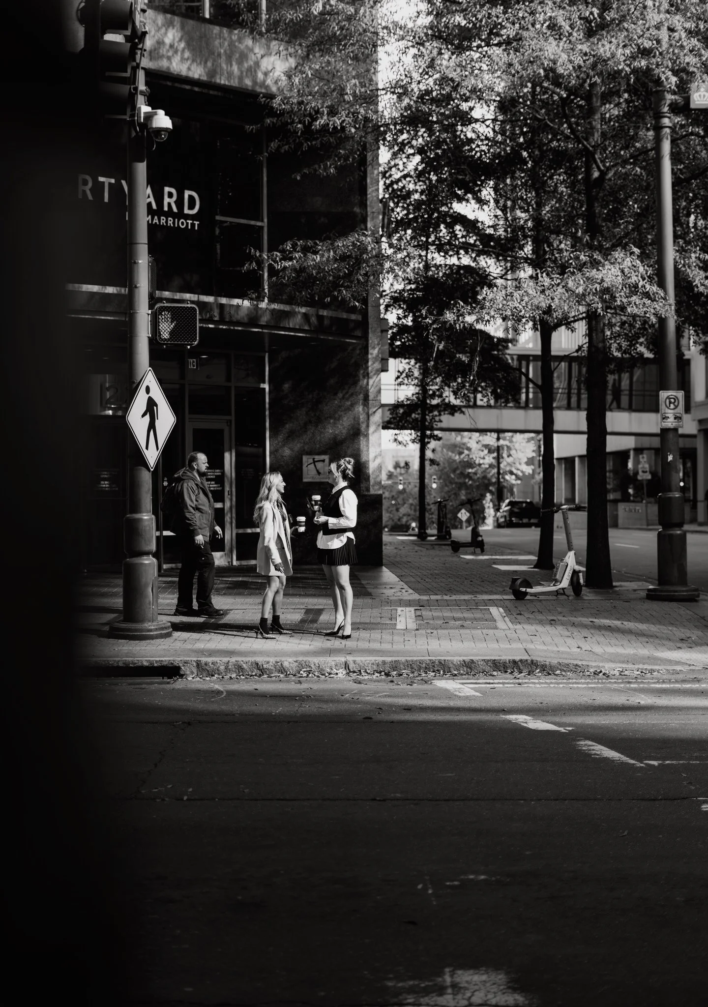 A city sidewalk scene in black and white showing three people, with two women and a man, standing and talking on the corner. One woman is holding a coffee cup, and the other woman and the man are listening. The background includes trees, a building with a sign that reads 'RTYARD MARRIOTT,' a scooter, and street signs, one indicating pedestrian crossing.