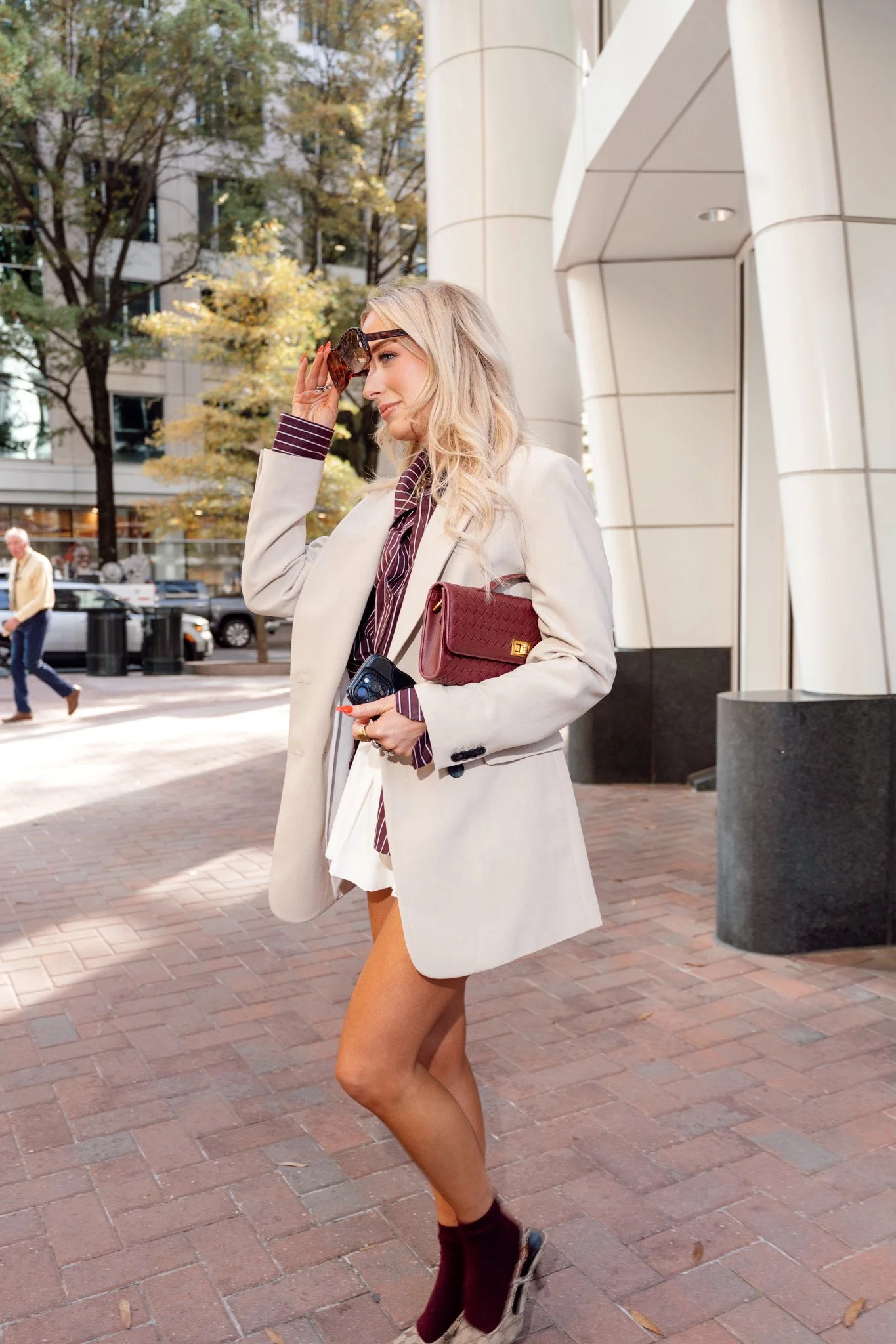 Woman walking outdoors in city, wearing a white blazer, striped shirt, shorts, maroon socks, sunglasses, and holding a maroon purse and glasses.