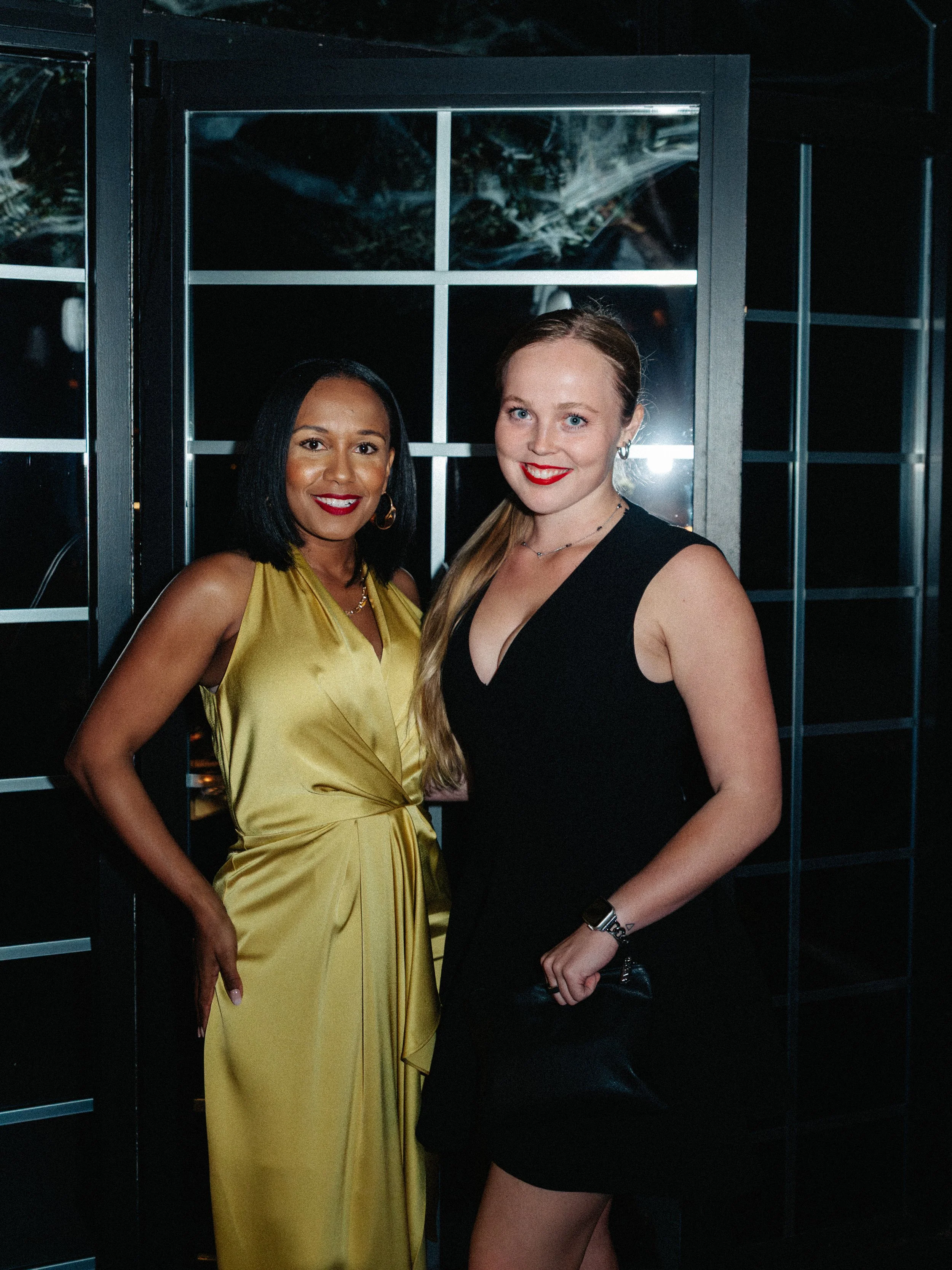 Two women dressed in evening wear posing together indoors at night.