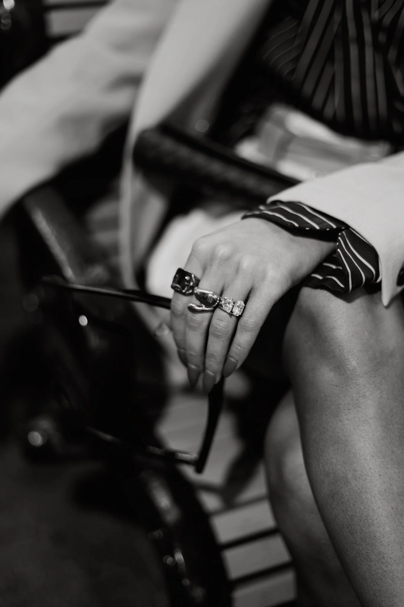 Close-up of a woman's hand with multiple rings resting on her knee, wearing a striped shirt and a light-colored blazer, sitting on a chair.
