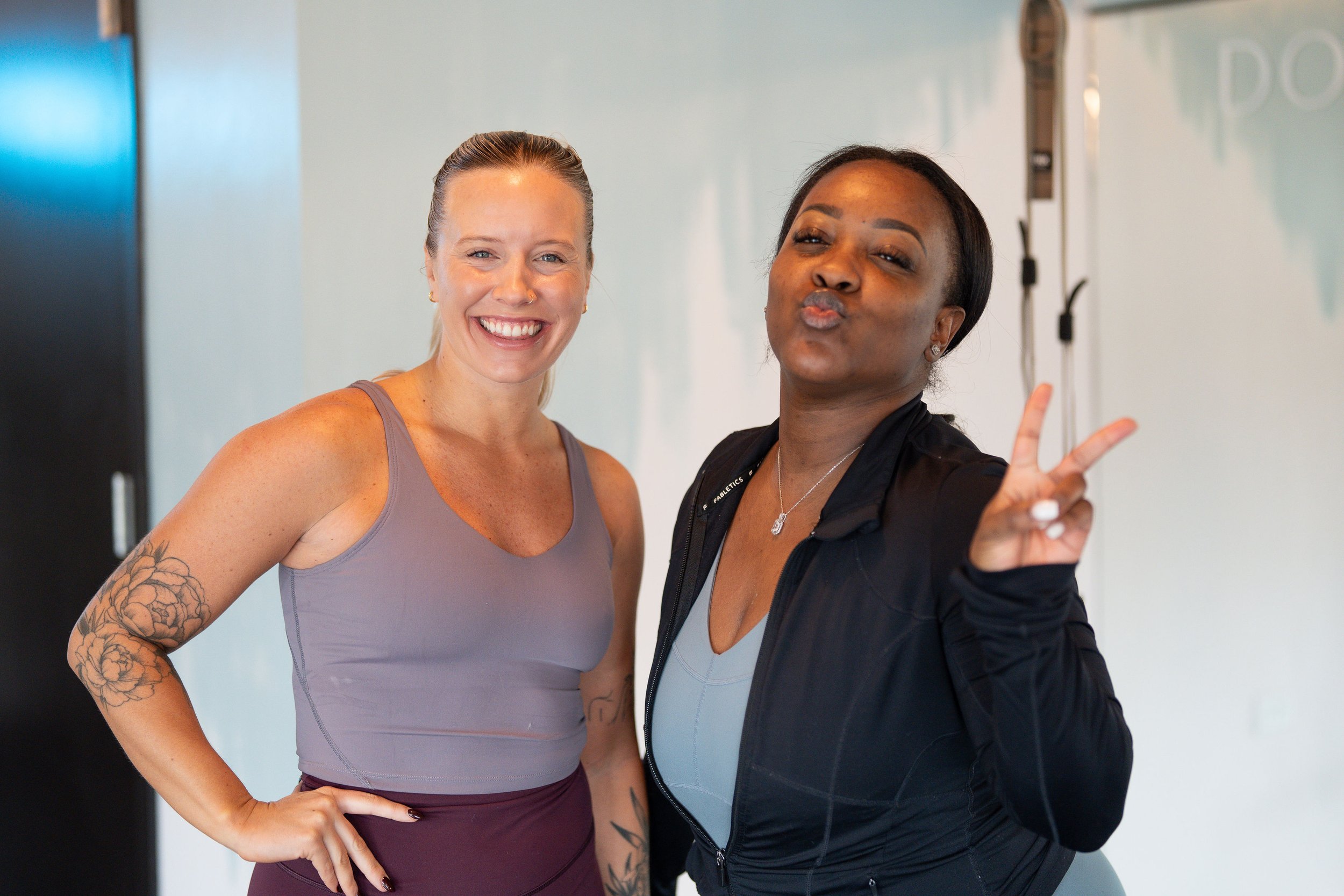Two women posing indoors, smiling and making peace signs. One has blonde hair, tattoos on her arm, and is wearing a gray workout top. The other has dark hair, is making a kiss face, and is wearing a black jacket over a fitness outfit.